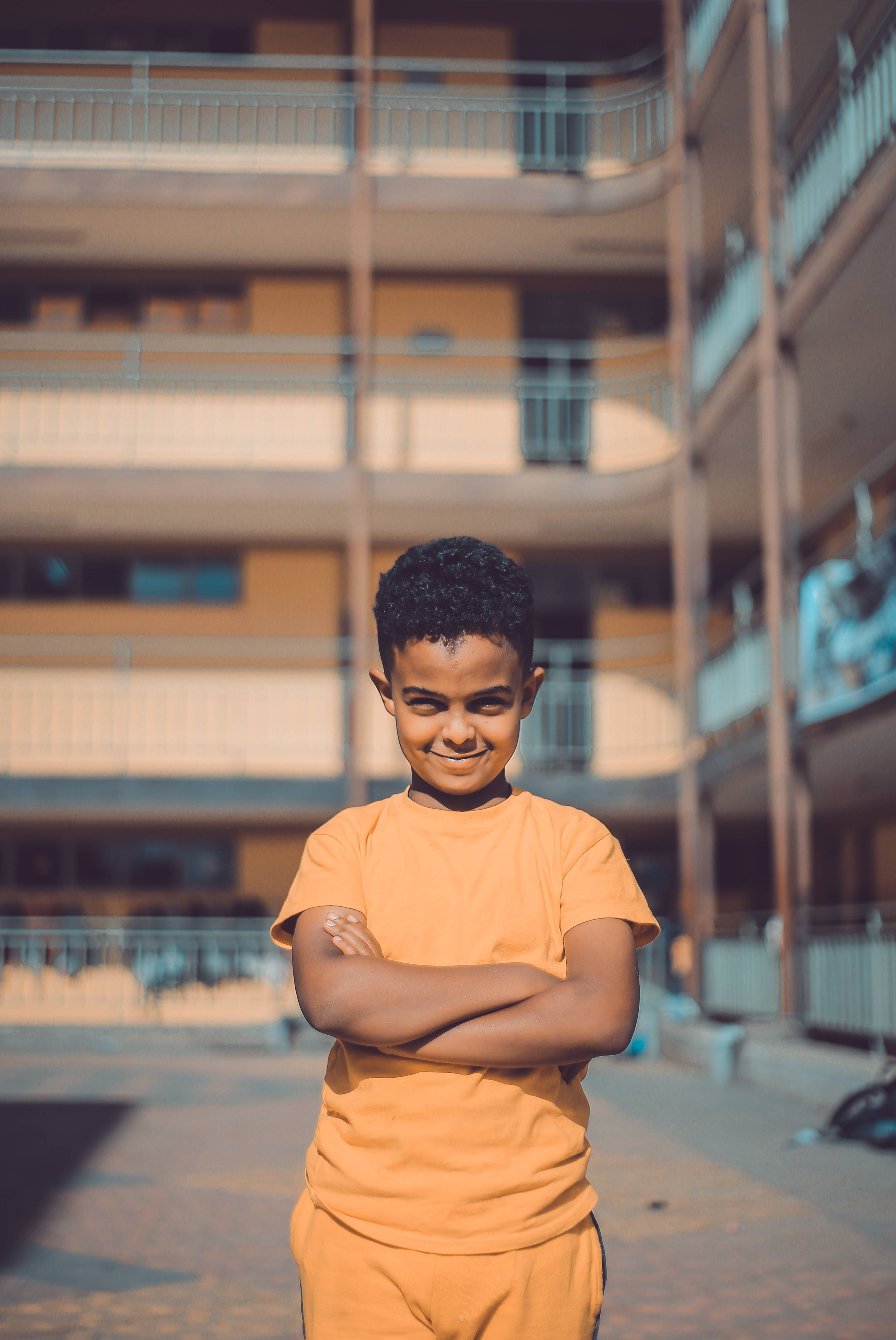 Ethiopia 桌布 壁紙 — 一個自信的男孩穿著橙色衣服，在公寓樓之間的陽光庭院中交叉雙臂站立。