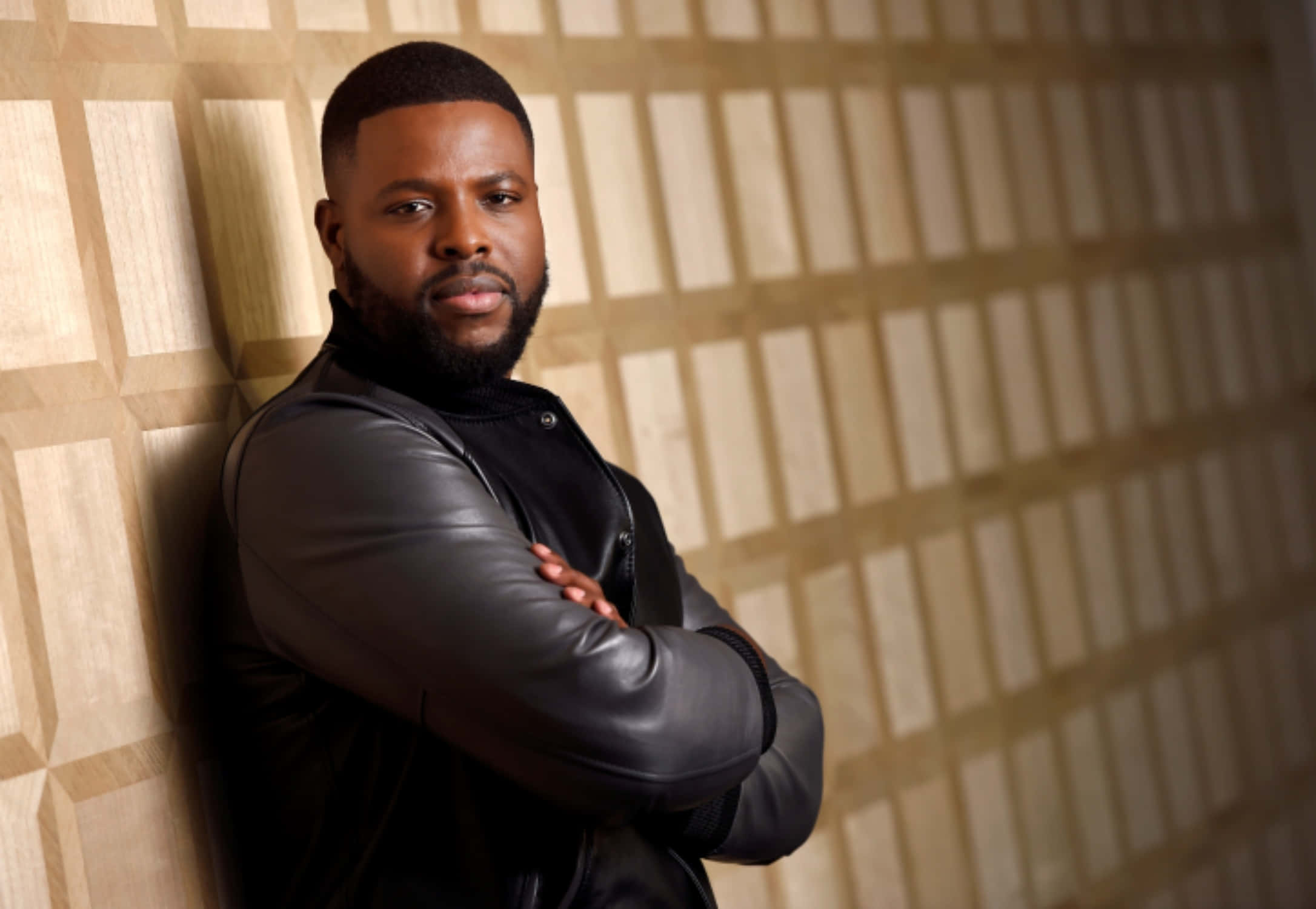 Winston Duke 桌布 壁紙 — 一個自信的鬍鬚男人靠在幾何圖案牆上，穿著黑色皮夾克。