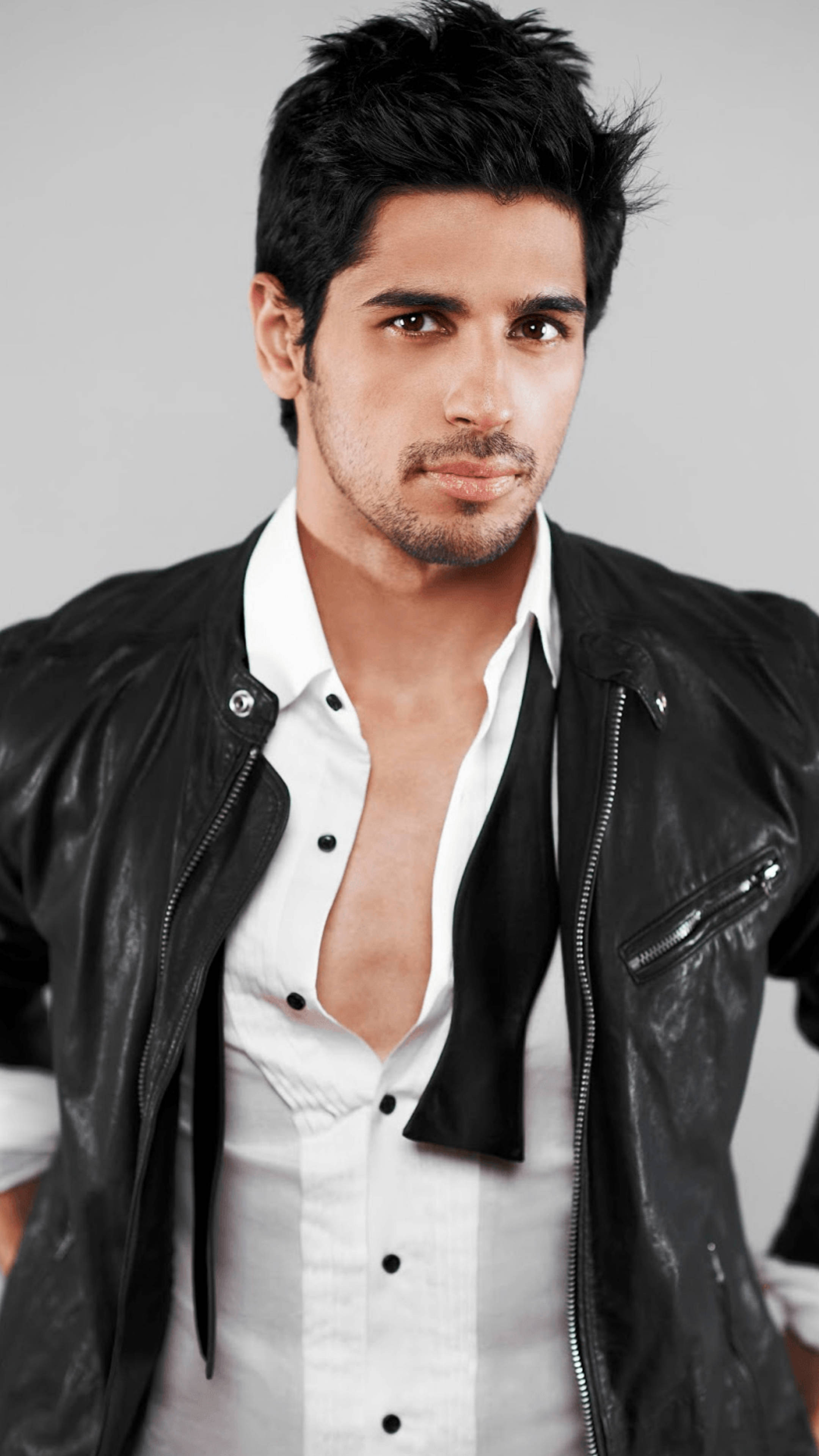 Sidharth Malhotra 桌布 壁紙 — 一個自信的黑髮鬍渣男人，身穿黑色皮夾克，內搭白色襯衫，鈕扣解開，背景是純灰色。