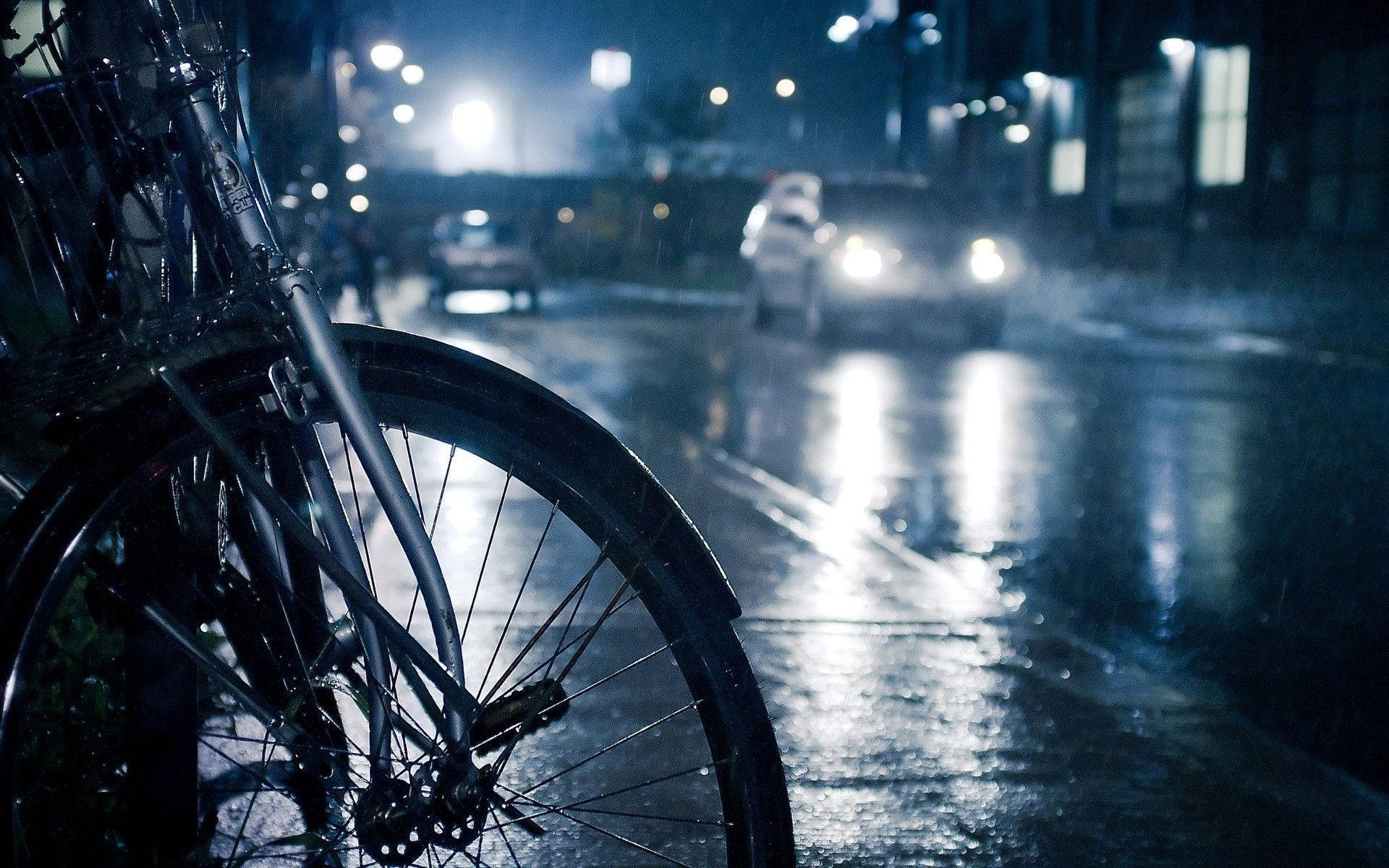 Raining 桌布 壁紙 — 一個自行車輪胎矗立在前景，雨濕的街道反射著夜晚的車頭燈。
