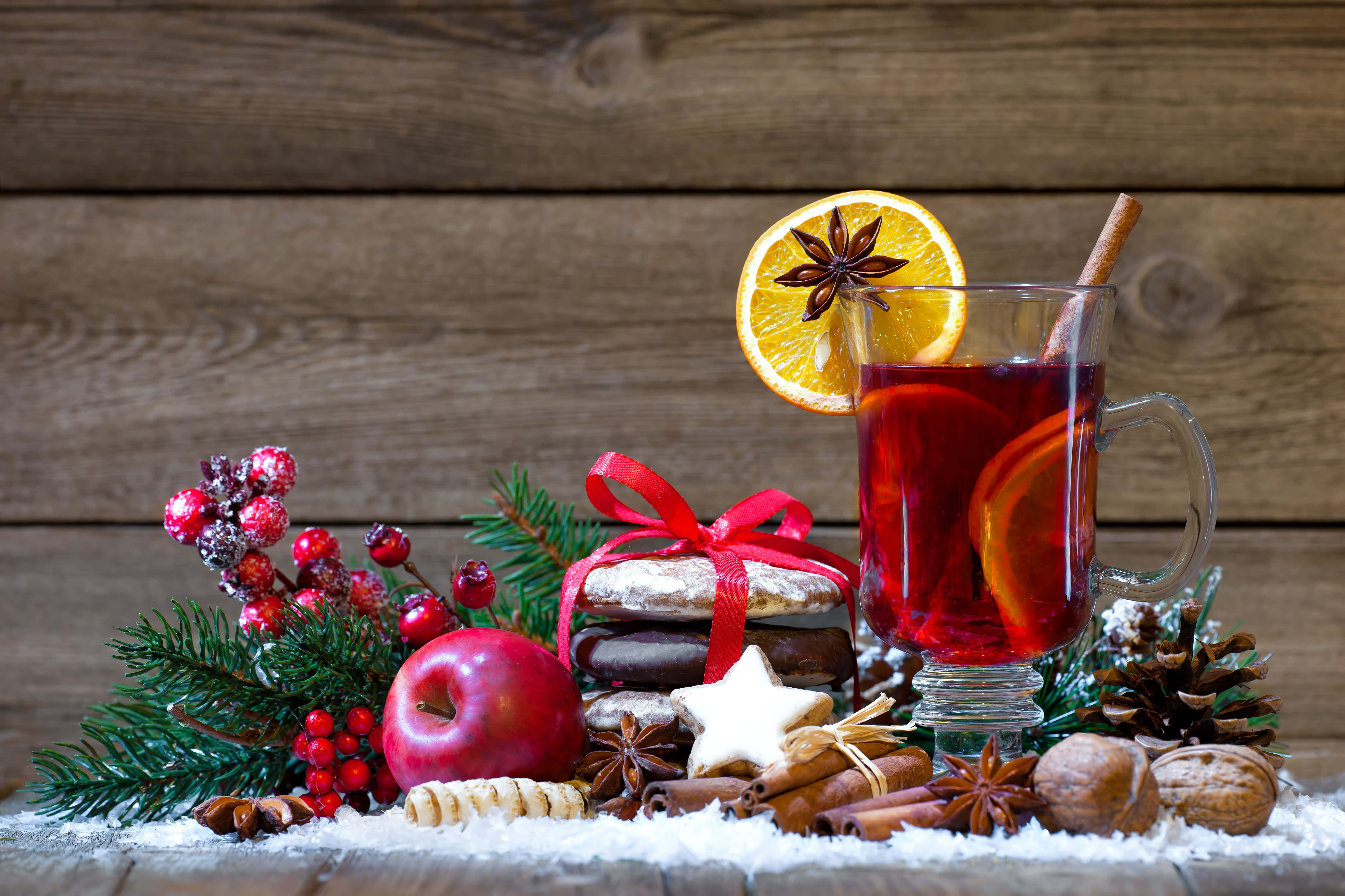 Mulled Wine 桌布 壁紙 — 一個舒適的冬季假日場景，一杯加了香料的热紅酒，以橙子和八角裝飾，旁邊有節日餅乾、松果和紅莓，放在雪覆蓋的木頭上。