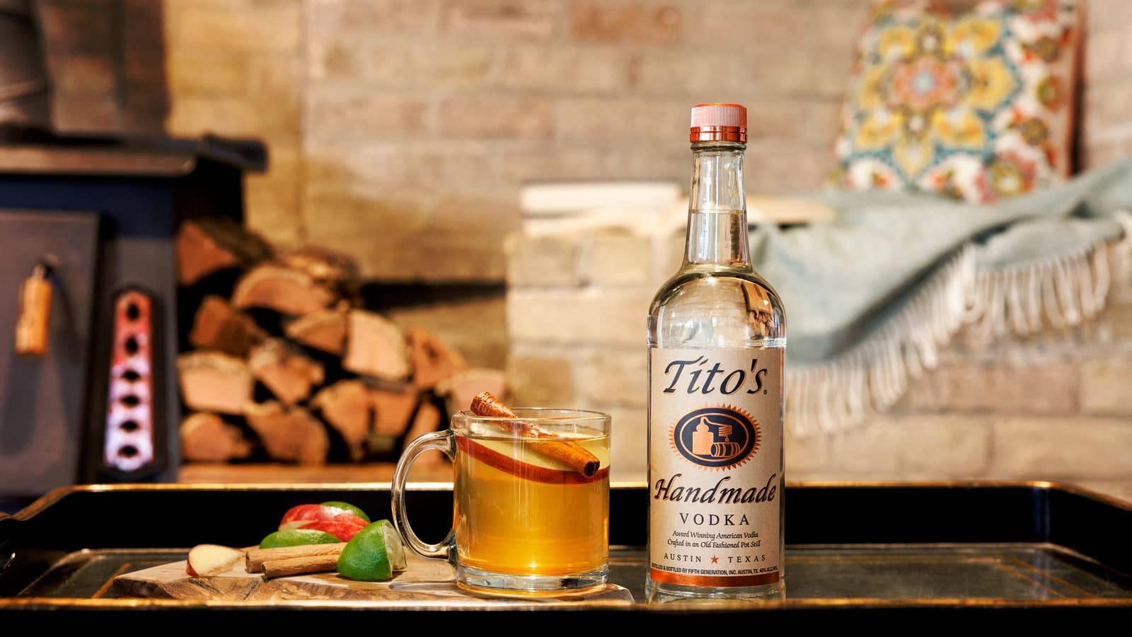 cocktail 桌布 壁紙 — 一個舒適的冬日場景，一杯香料蘋果酒和Tito's伏特加放在壁爐旁，壁爐裡堆滿了木柴。