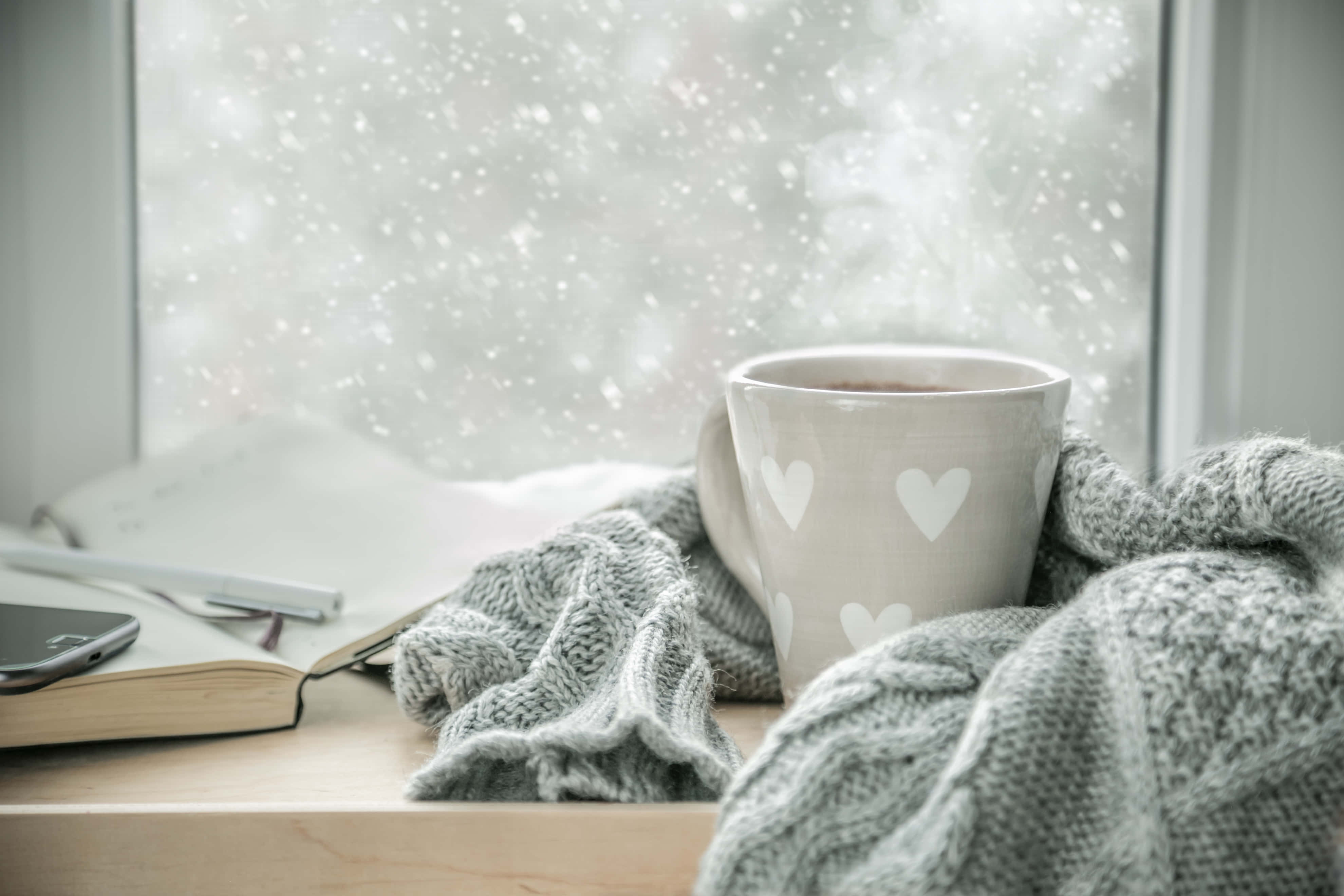 Hot Chocolate 桌布 壁紙 — 一個舒適的冬日場景，窗台上放著一個冒著熱氣的心形圖案馬克杯，裹在針織毯子裡，旁邊有一本打開的筆記本，窗外雪花飄落。