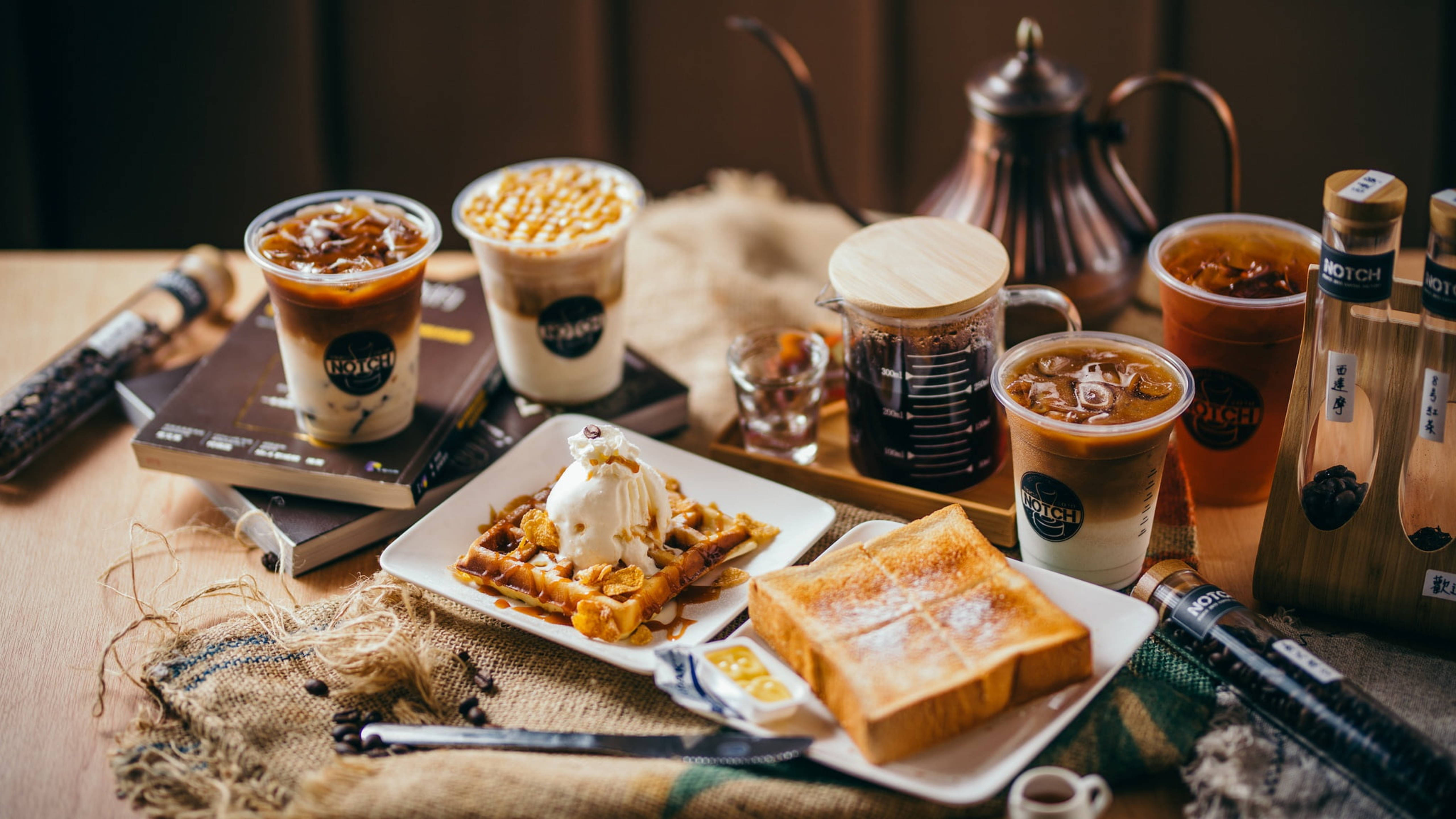 Waffle 桌布 壁紙 — 一個舒適的咖啡店擺設，包括冰咖啡飲品、鬆餅、吐司和書籍，整齊排列在木桌上，配以溫暖燈光。