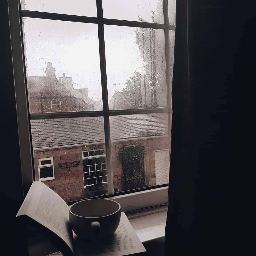 Dark Academia Aesthetic 桌布 壁紙 — 一個舒適的室內場景，窗台上放著一本打開的書和一個杯子，雨滴在玻璃上劃出條紋，而柔和的磚建築在灰色的天空中逐漸消失。
