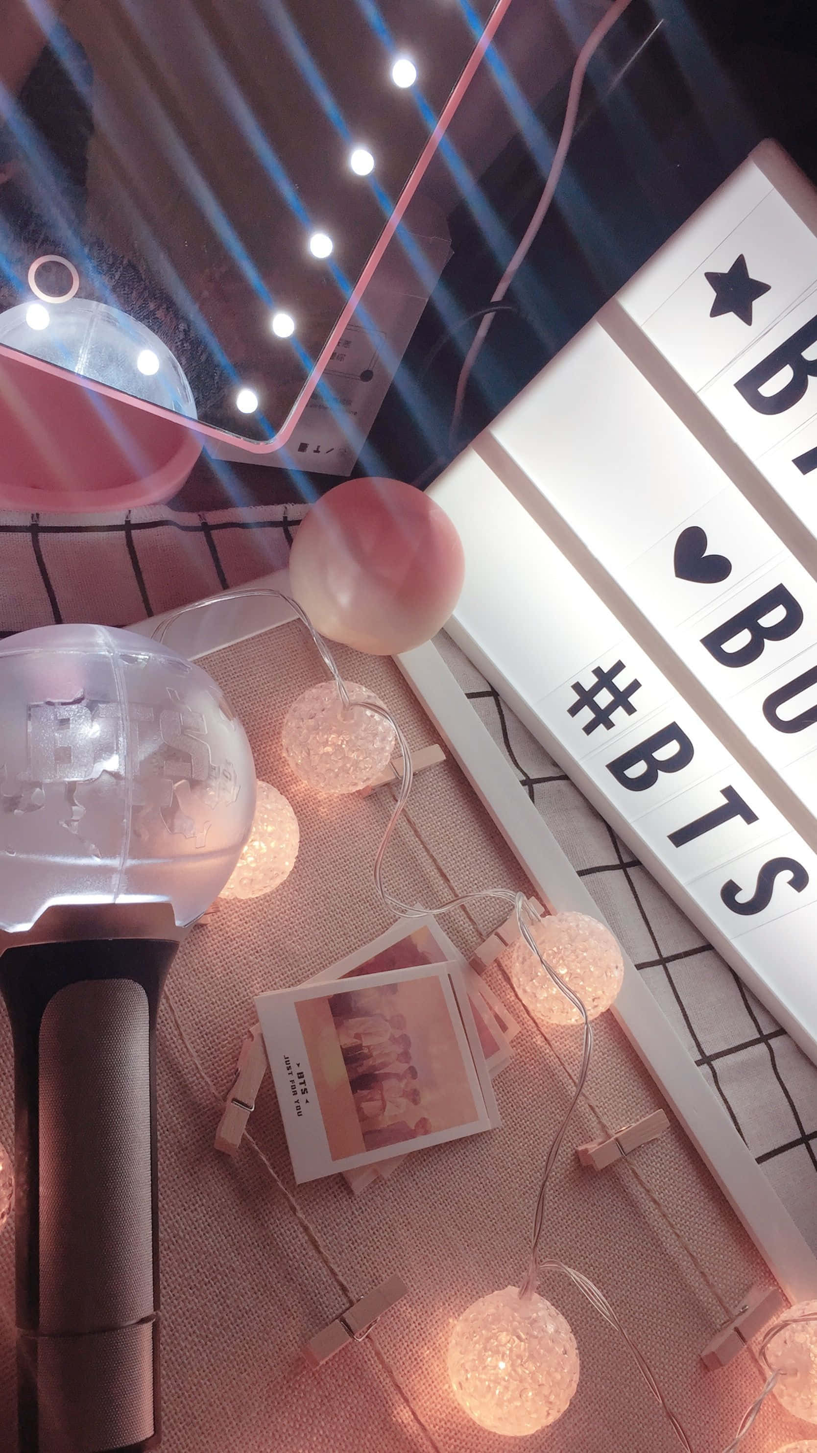 Bts Army Bomb 桌布 壁紙 — 一個舒適的室內場景，粉紅色串燈在BTS燈箱和拍立得照片旁邊溫暖地發光，放在格子表面上。