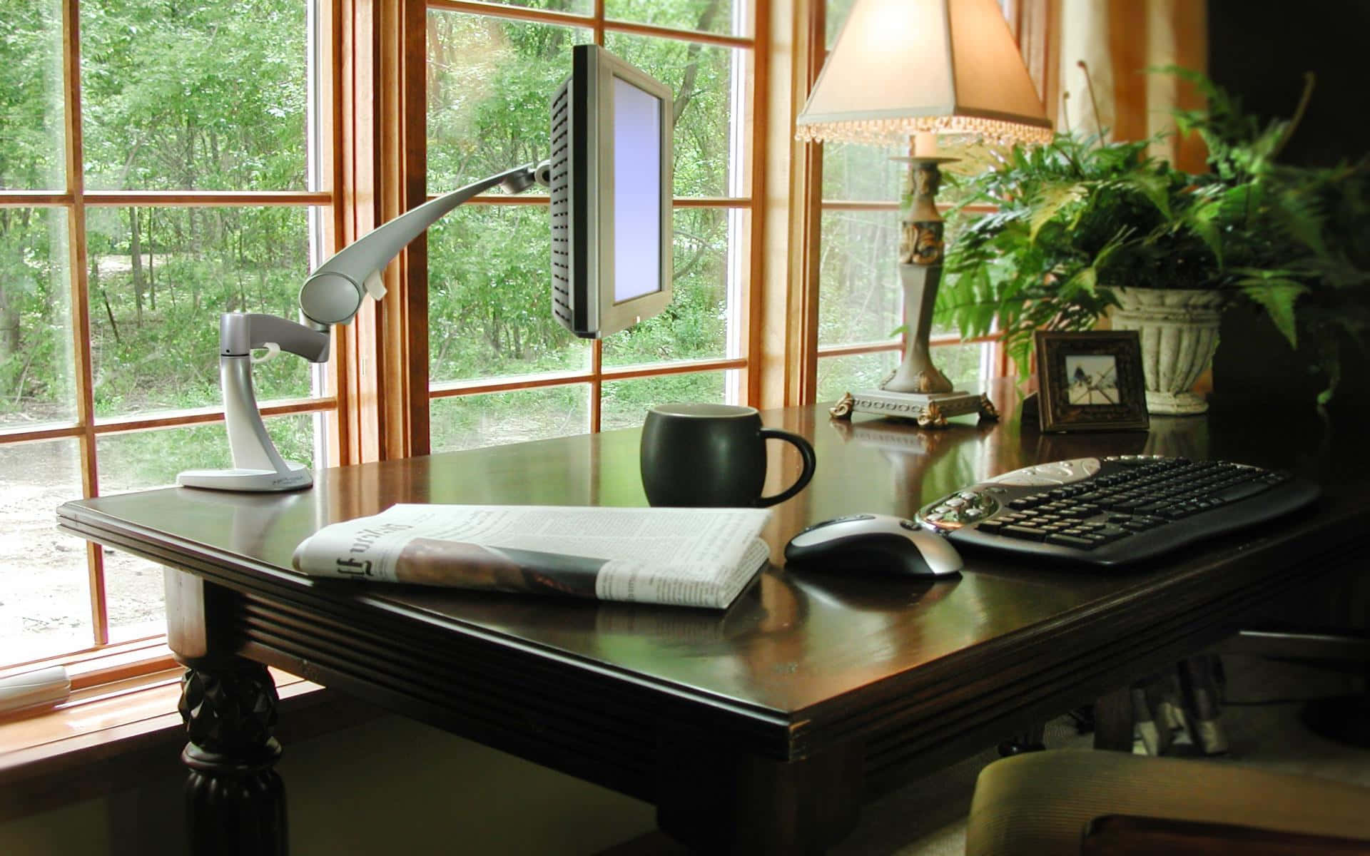 Cozy Desktop 桌布 壁紙 — 一個舒適的家居辦公室書桌，靠近大窗戶，窗外有綠樹，配備顯示器燈、馬克杯、鍵盤和植物。