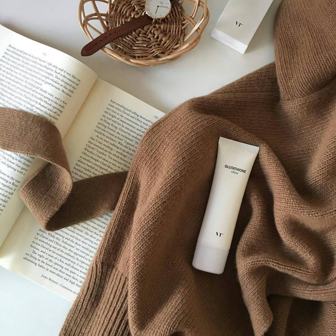 Beige Minimalist 桌布 壁紙 — 一個舒適的平鋪構圖，包含棕色針織毛衣、打開的書本、編織籃內的手錶，以及白色穀胱甘肽面霜，置於白色表面上。