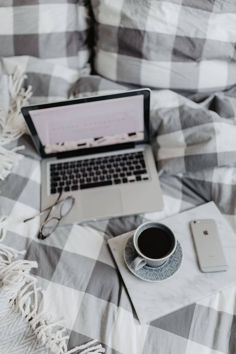 咖啡動圖 桌布 壁紙 — 一個舒適的床鋪設置，在柔和自然光下，格子毯上放有筆記型電腦、咖啡杯和手機。