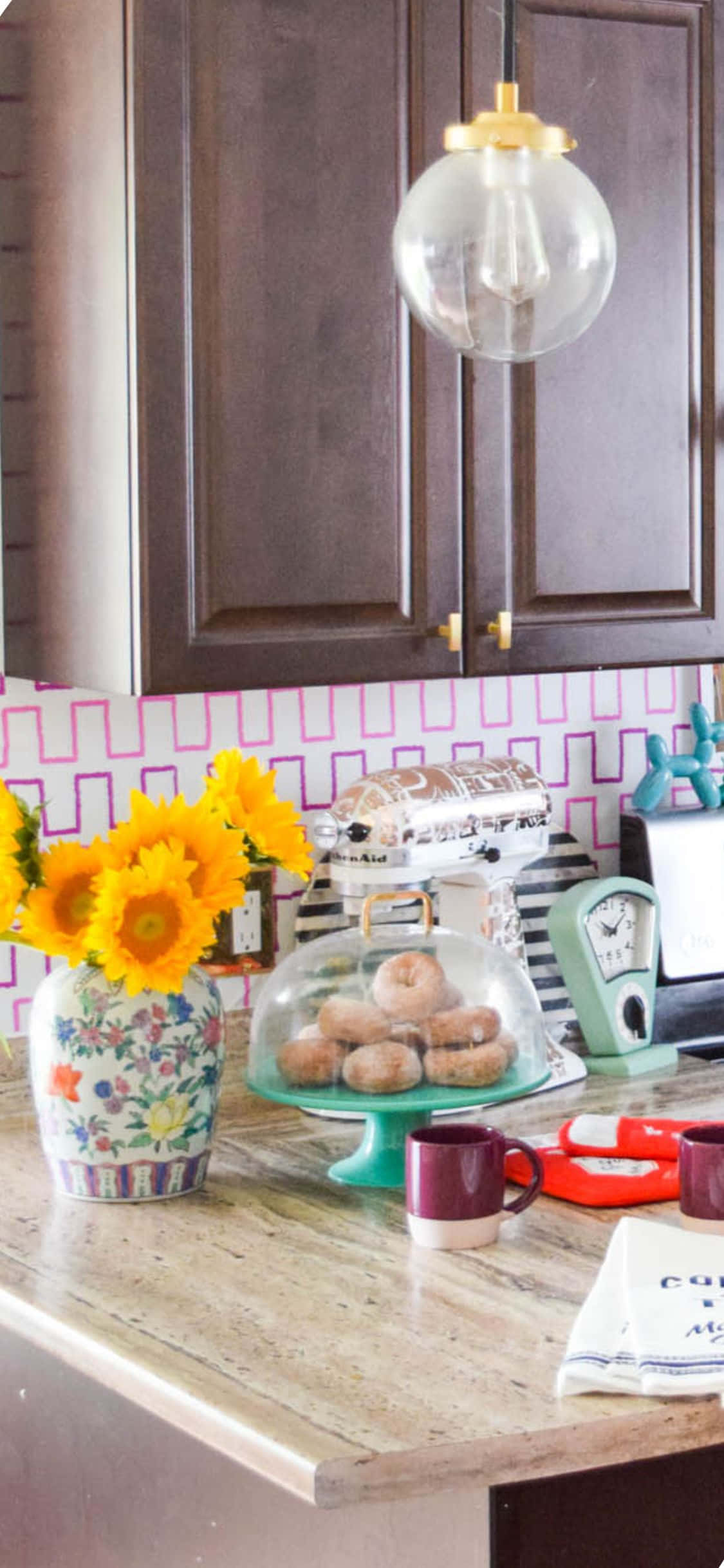 Kitchen Design 桌布 壁紙 — 一個舒適的廚房檯面，擺放著花卉花瓶中的向日葵、玻璃罩下的甜甜圈，以及復古電器，背景是粉紅色幾何圖案防濺板。