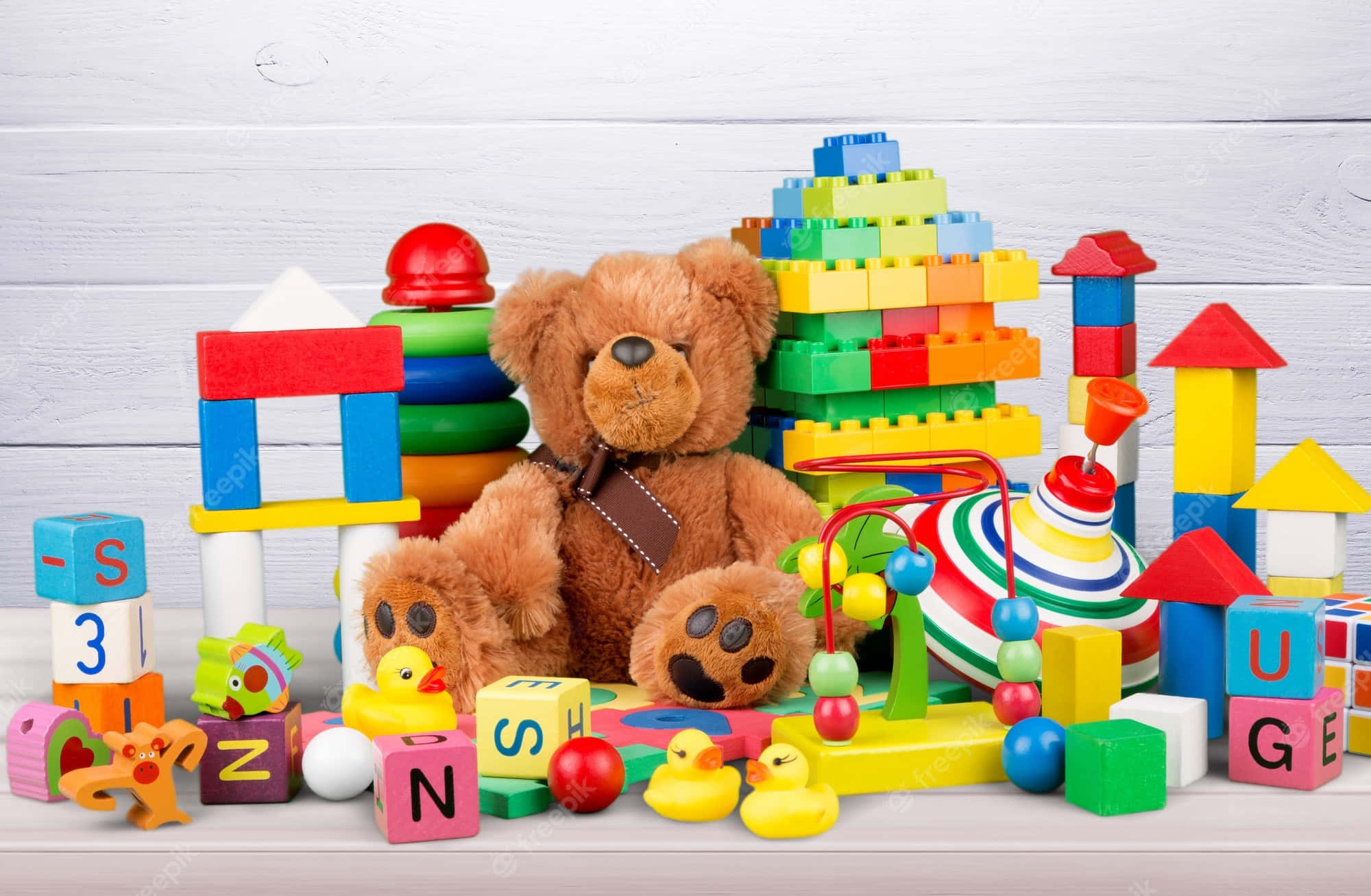 toys 桌布 壁紙 — 一個舒適的彩色兒童玩具系列，包括一隻棕色泰迪熊，周圍環繞著字母積木和塑膠建築積木，放在白色木質表面上。