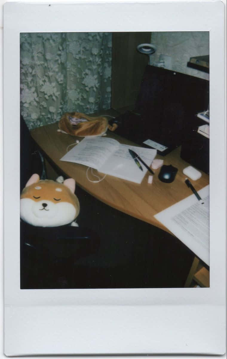 Aesthetic Polaroid 桌布 壁紙 — 一個舒適的書桌設置，椅子上有一個毛絨柴犬頭，紙張、筆和筆記型電腦在溫暖的室內燈光下。