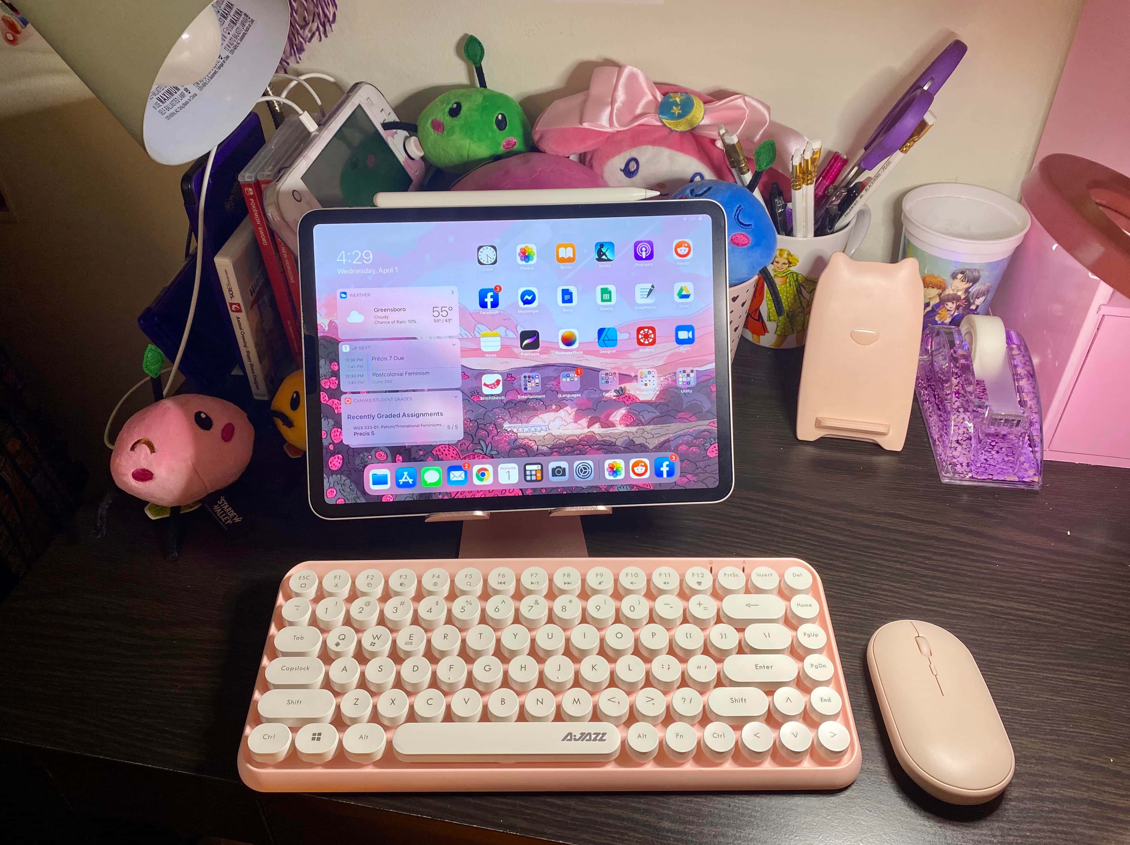 美學 iPad 壁紙 桌布 壁紙 — 一個舒適的桌面設置，配有粉紅色鍵盤、滑鼠和平板電腦，顯示充滿活力的日落桌布，周圍環繞著毛絨玩具和文具。