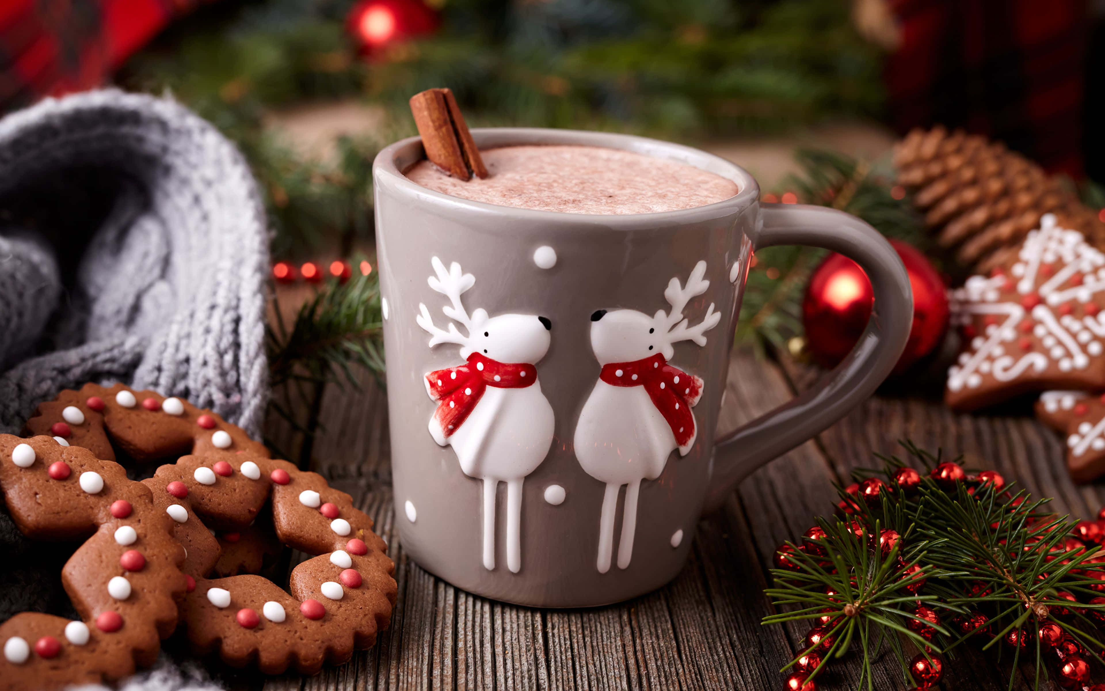 Hot Chocolate 桌布 壁紙 — 一個舒適的灰色馴鹿圖案馬克杯，盛載著香料熱巧克力，上面放有肉桂棒，周圍有薑餅餅乾、松果和節日紅色裝飾，放在鄉村風木桌上。