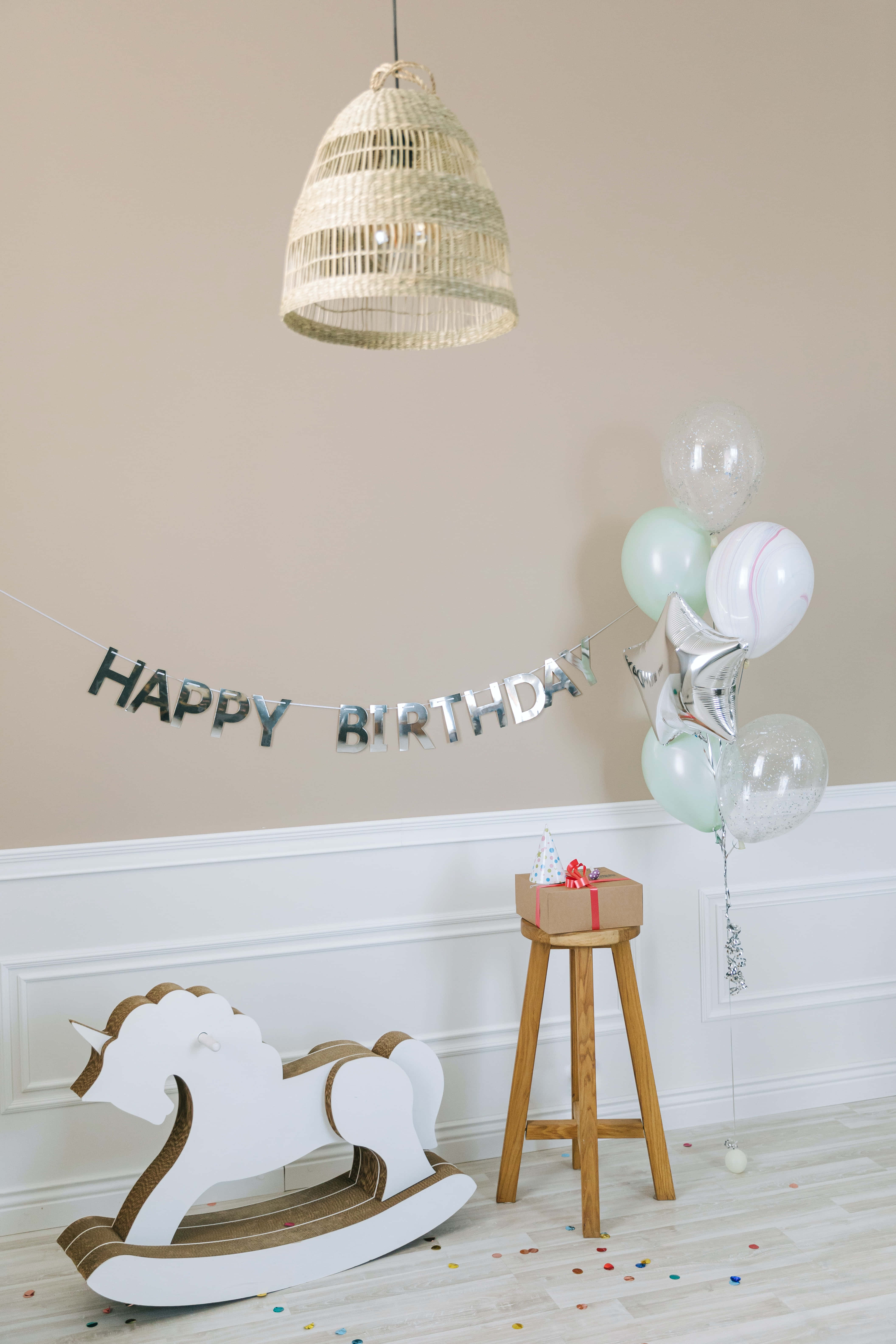 Cute Birthday 桌布 壁紙 — 一個舒適的生日佈置，包含白色搖馬、銀色氣球，以及編織吊燈下的「生日快樂」橫幅。