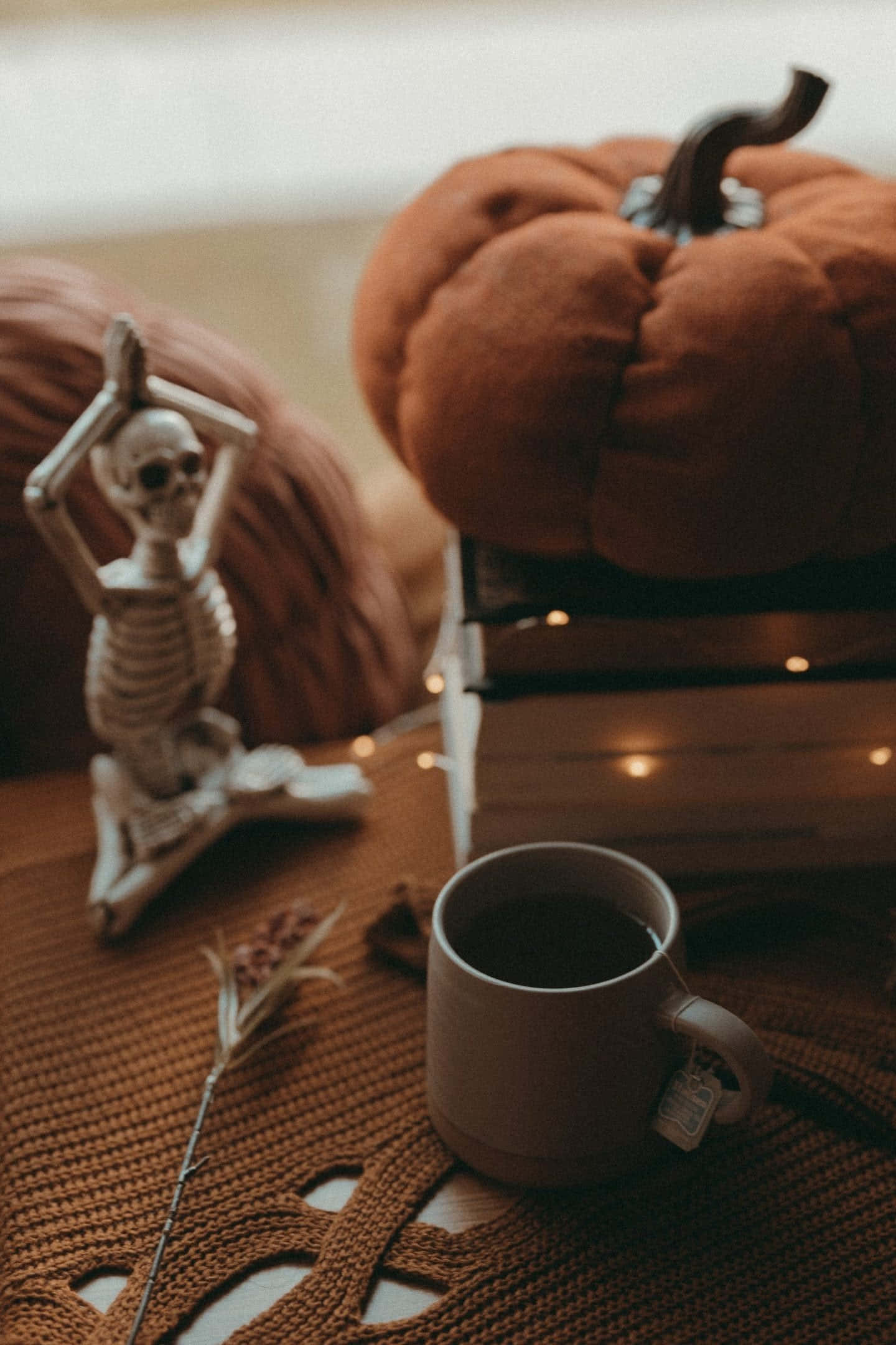 Fall Halloween iPhone 桌布 壁紙 — 一個舒適的秋季場景，一杯茶旁邊有一個毛絨南瓜和一個瑜伽骷髏公仔放在針織毯上。