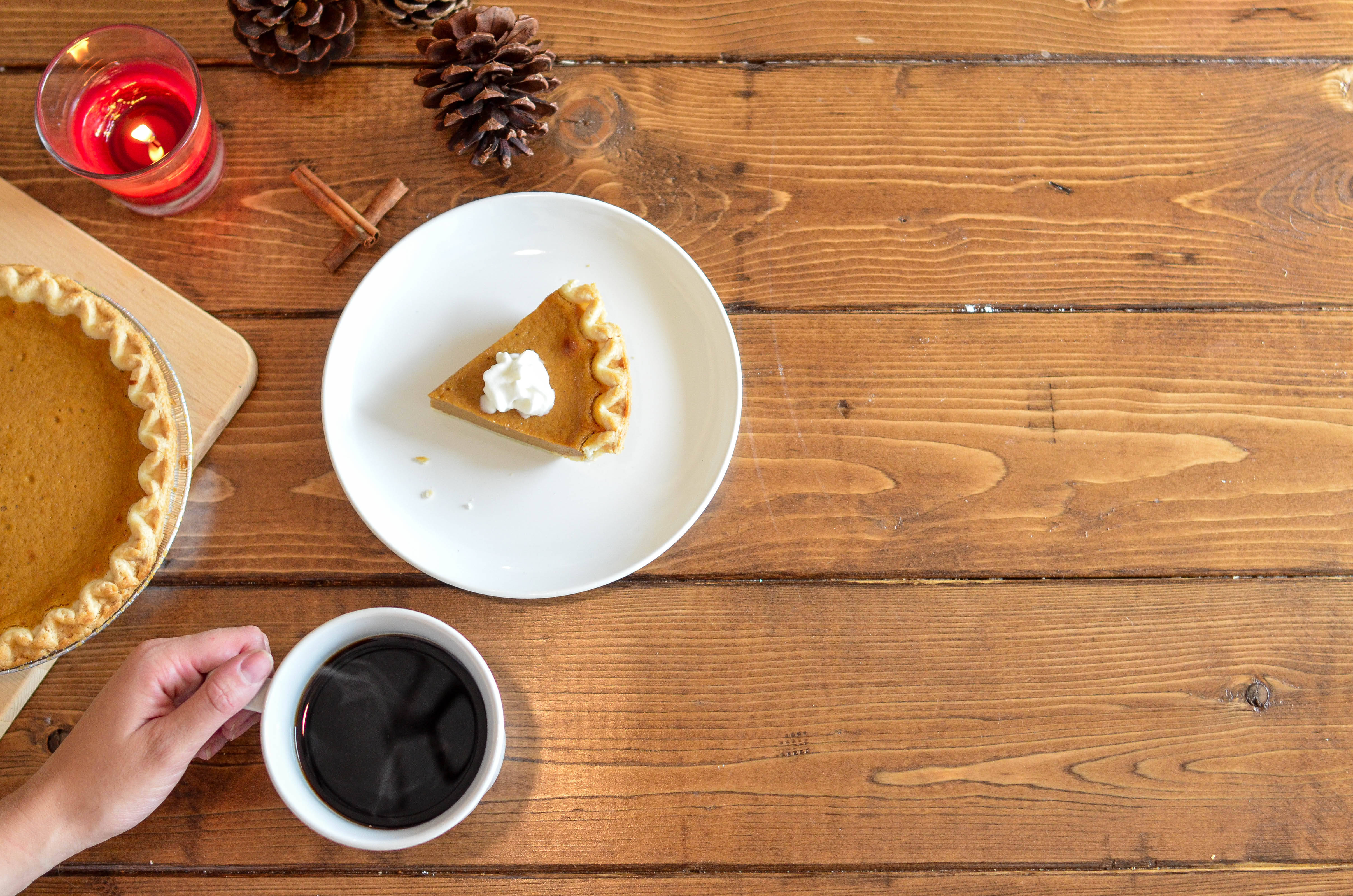 Happy Thanksgiving 桌布 壁紙 — 一個舒適的秋季場景，一片南瓜派上放有鮮奶油，放在白色盤子上，旁邊有一隻手拿著一杯咖啡和一支點燃的蠟燭，放在質樸的木桌上。
