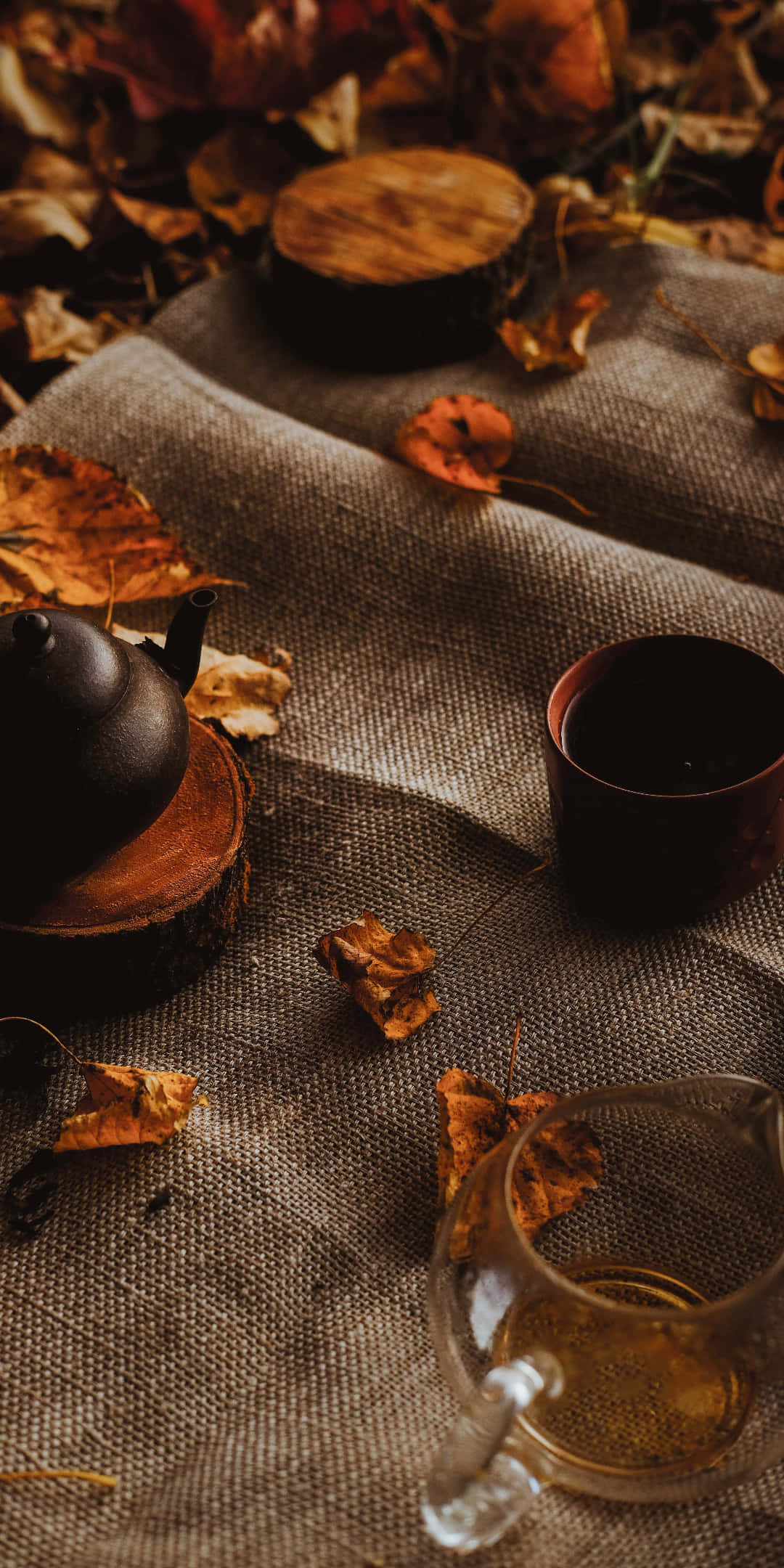 Pixel 3 Autumn 桌布 壁紙 — 一個舒適的秋季茶具擺設，黑色茶壺和玻璃壺放在麻布上，周圍散落著落葉。