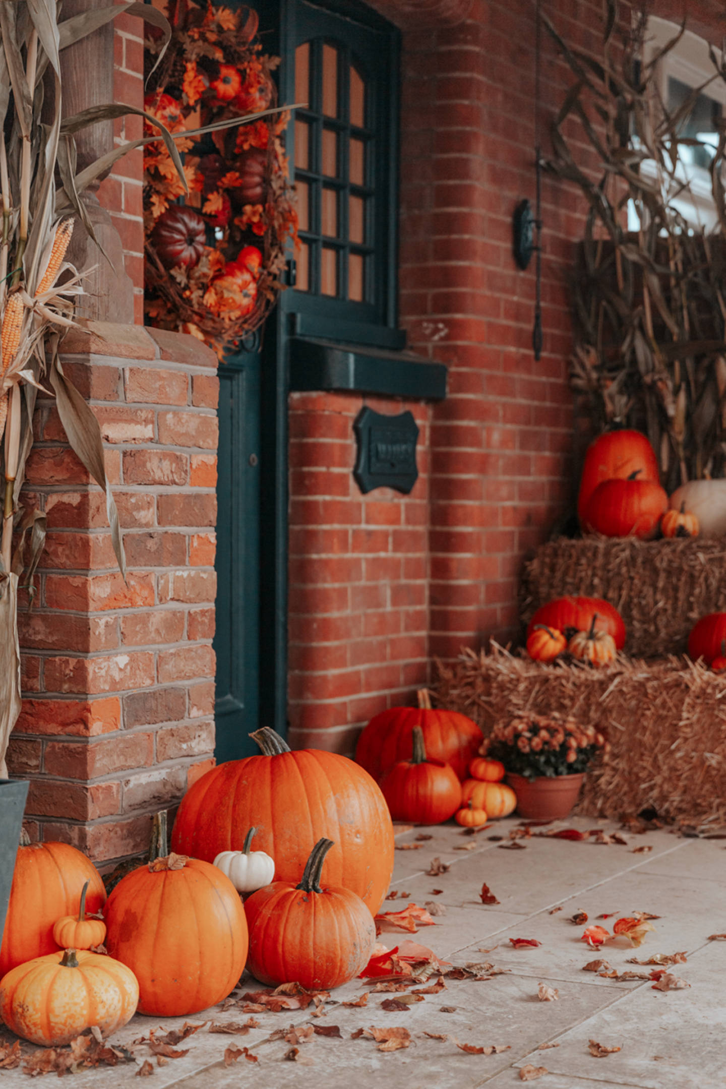 Halloween Aesthetic 桌布 壁紙 — 一個舒適的秋季門廊，裝飾著南瓜、玉米稈和花環，背景是磚牆和深綠色門。