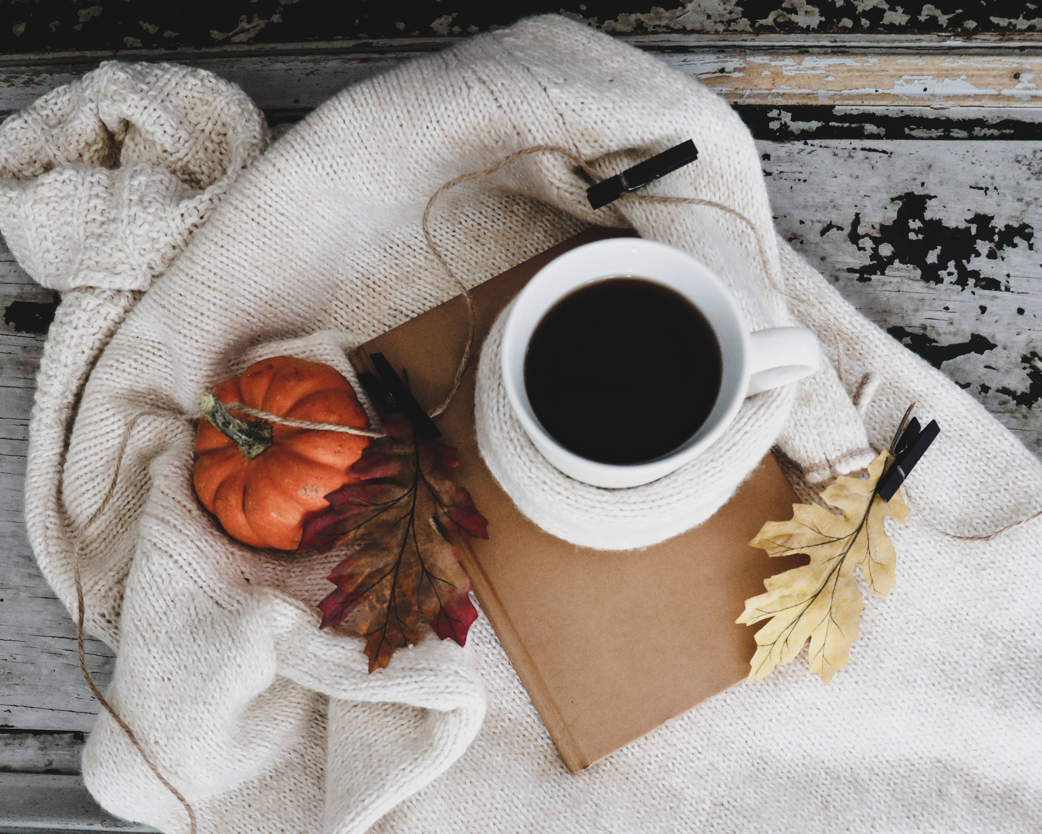 Fall 桌布 壁紙 — 一個舒適的秋日場景，一杯白色咖啡放在書本上，書本擱在米色針織毛衣上，周圍有南瓜和彩色樹葉。