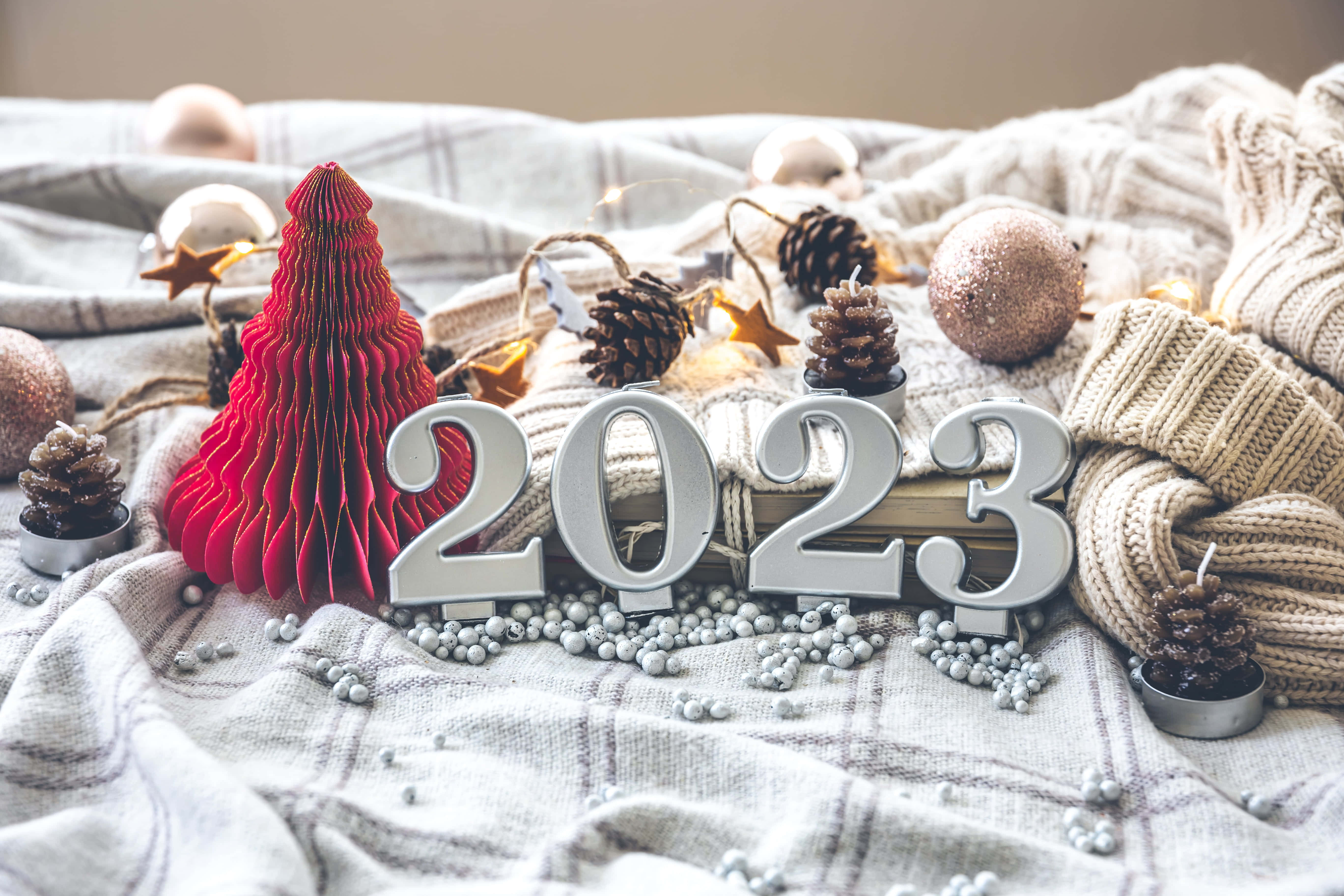 New Year 桌布 壁紙 — 一個舒適的節日場景，有銀色2023數字、紅色紙樹、松果和針織品放在格子毯上。