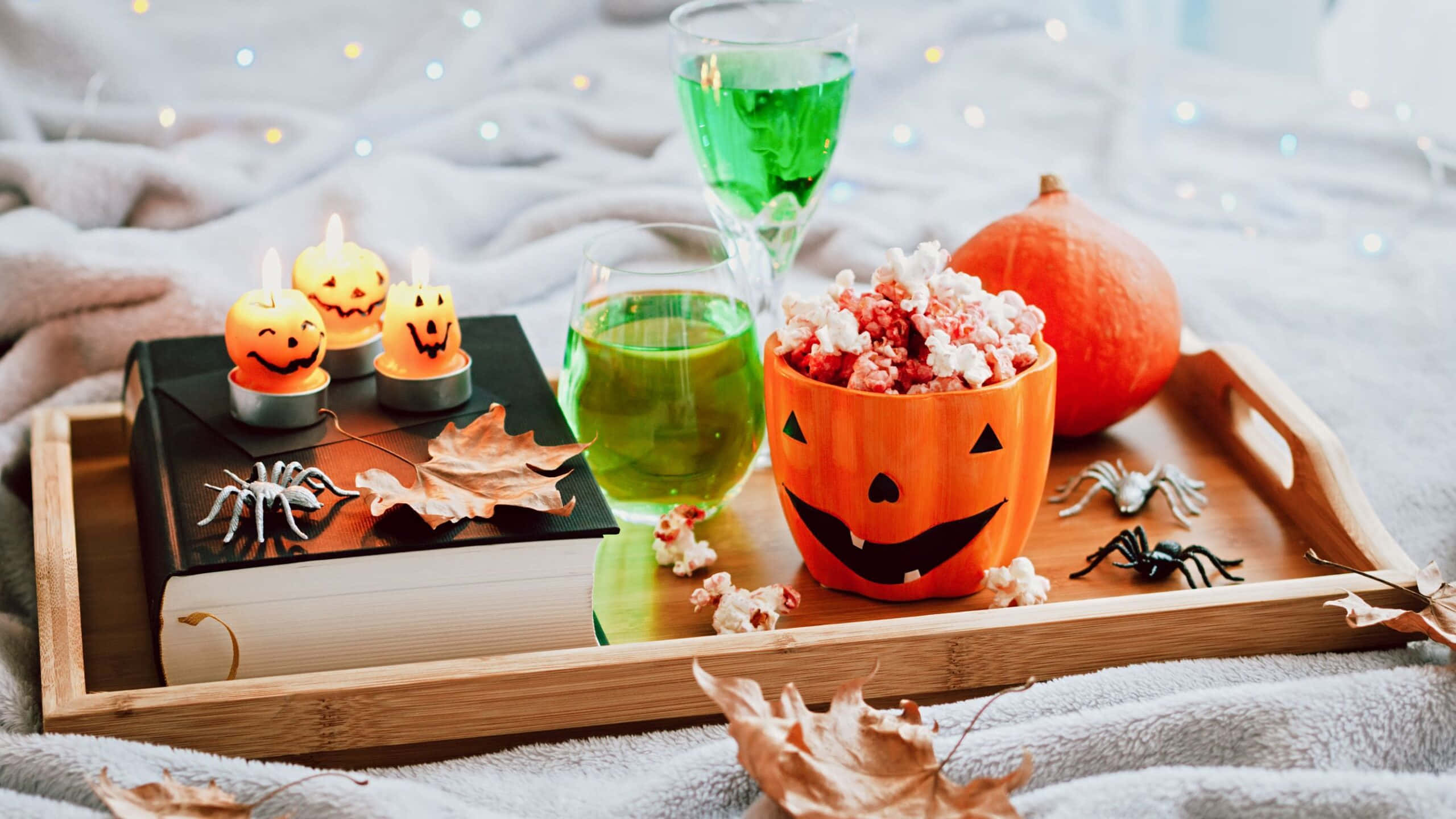 Halloween Cocktails 桌布 壁紙 — 一個舒適的萬聖節托盤，上面有南瓜爆米花、綠色飲料、傑克南瓜燈蠟燭和假蜘蛛，放在帶有仙女燈的床上。