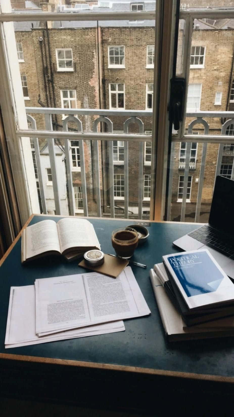 Studying 桌布 壁紙 — 一個舒適的讀書角落，暗色書桌上放著打開的書籍、紙張和一杯咖啡，透過玻璃門俯瞰磚砌建築。