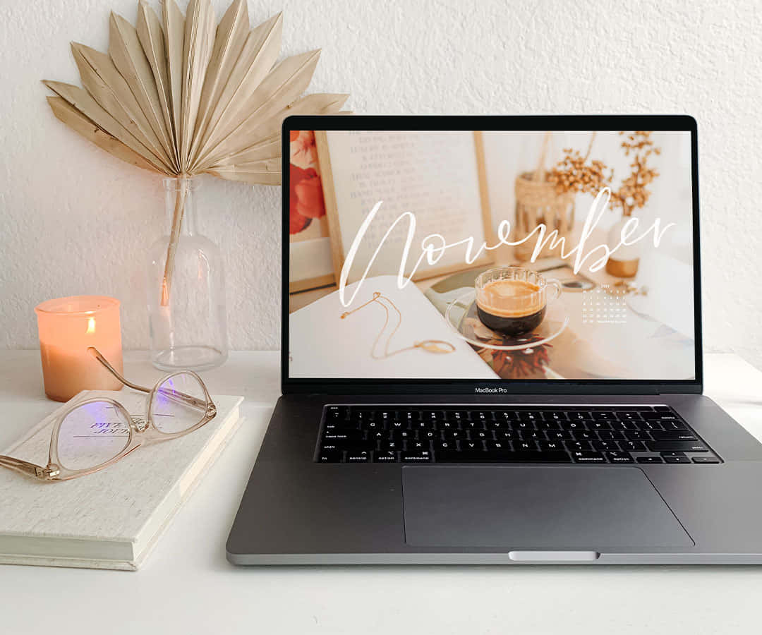 MacBook 動態圖片 桌布 壁紙 — 一個舒適的辦公桌設置，包括點燃的蠟燭、眼鏡和一部顯示十一月主題桌布的筆記型電腦，桌布上有咖啡和乾花。