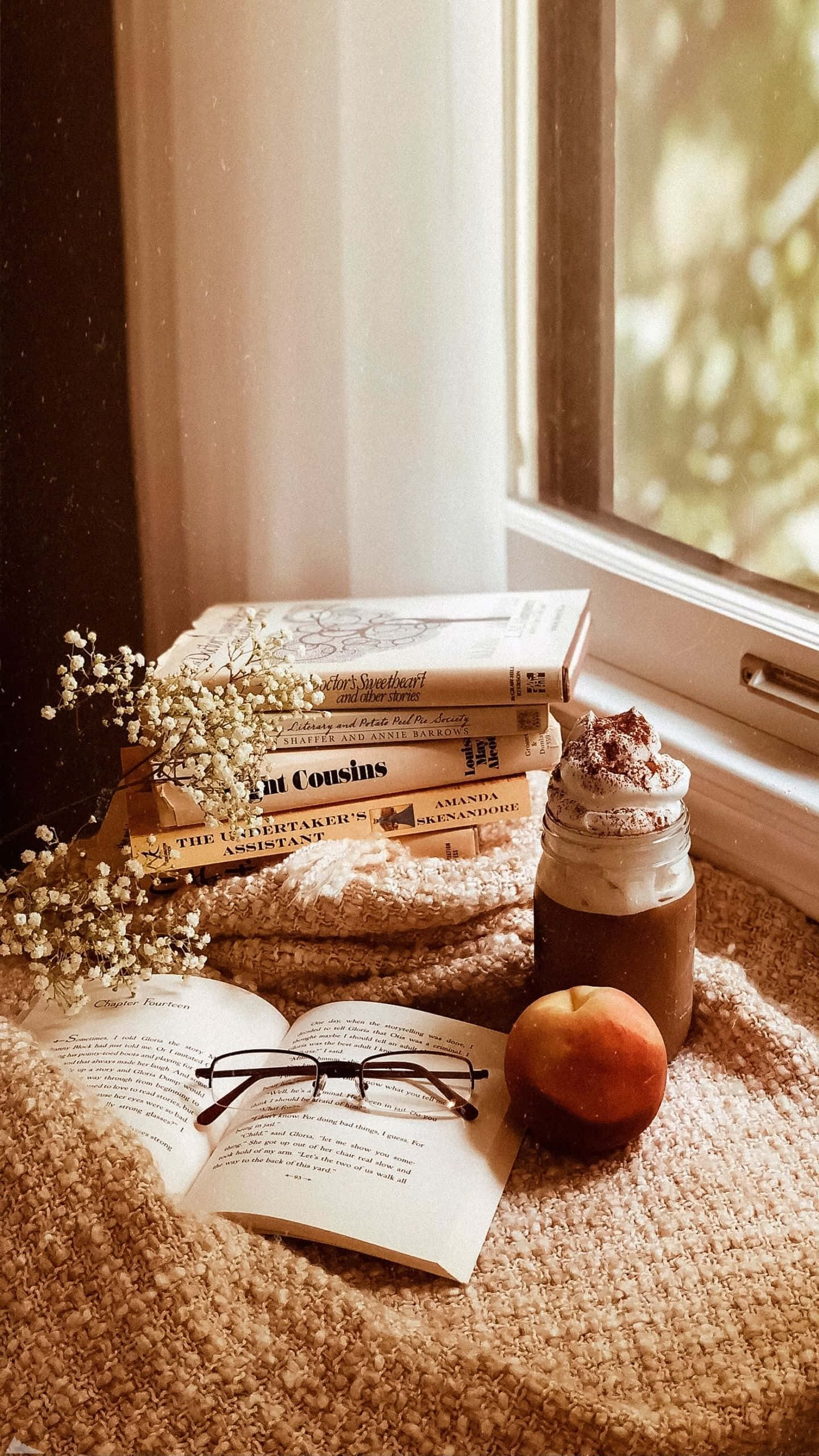 Cute Autumn iPhone 桌布 壁紙 — 一個舒適的閱讀角落，位於陽光充足的窗邊，有堆疊的書籍、咖啡杯和一個放在質感毛毯上的桃子。