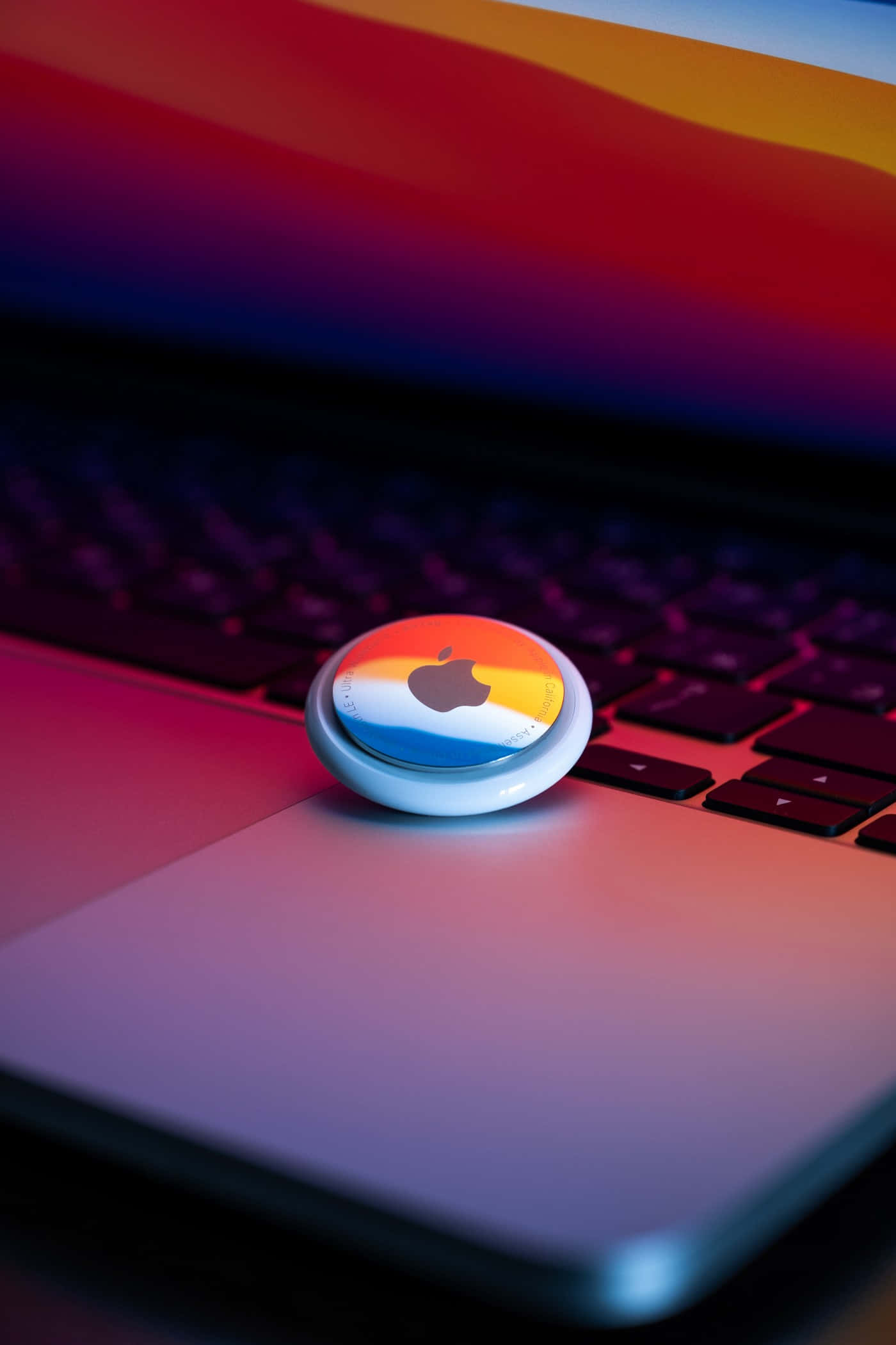 Pixel 3 Apple 桌布 壁紙 — 一個色彩繽紛的Apple AirTag放在一個沐浴在鮮豔漸變燈光下的筆記型電腦鍵盤上。