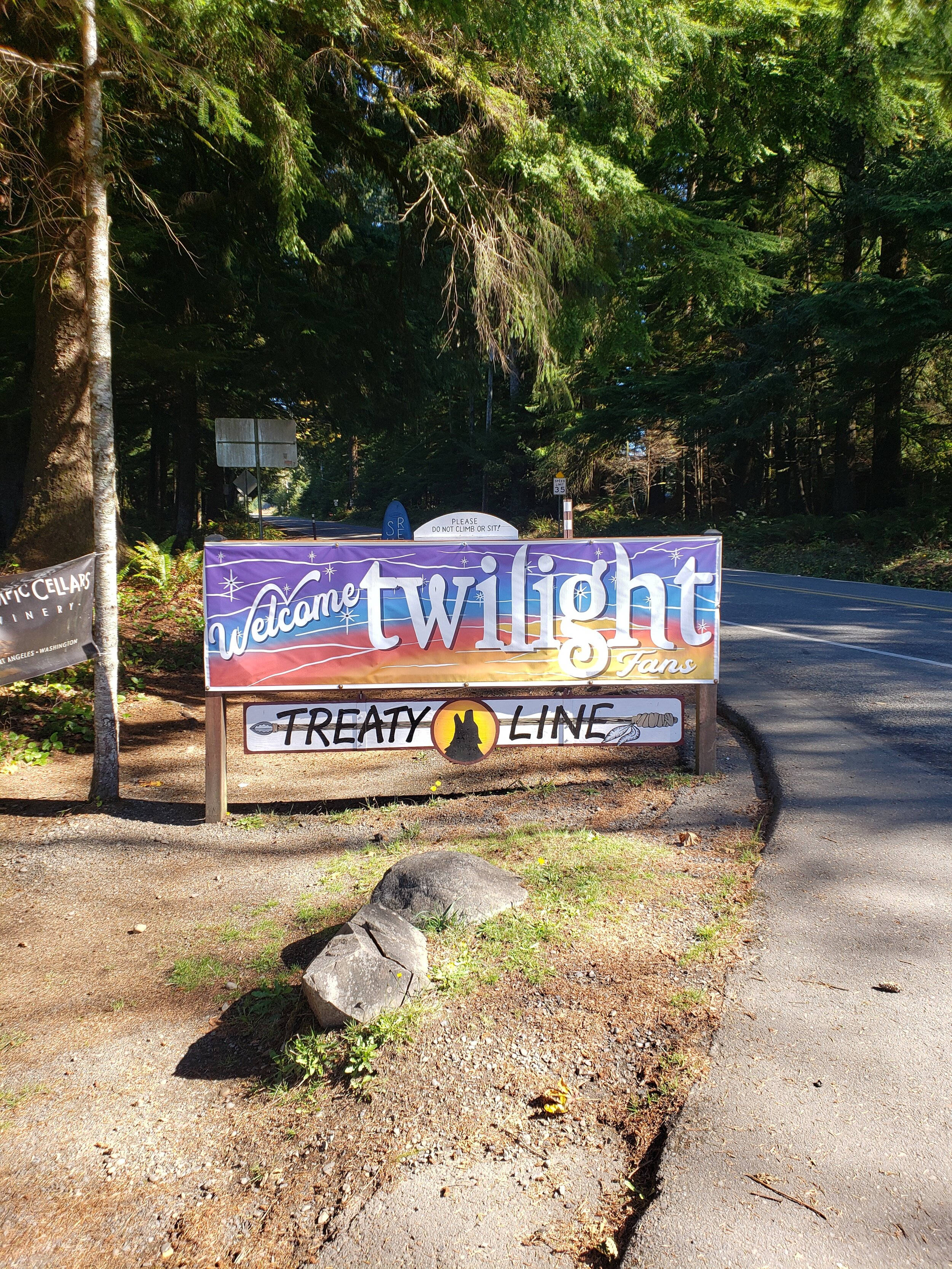 Forks 桌布 壁紙 — 一個色彩繽紛的Twilight粉絲歡迎牌豎立在樹林區的路邊，周圍有高大的樹木和一片碎石地。