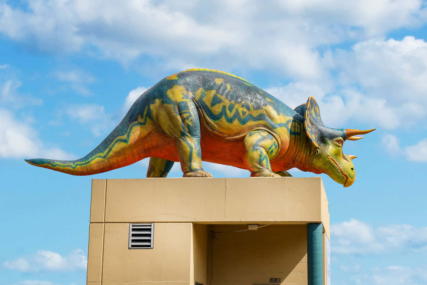Dino 桌布 壁紙 — 一個色彩繽紛的三角龍雕像，帶有藍色、黃色和橙色圖案，矗立在一棟米色建築物頂上，背景是明亮的藍天和蓬鬆的白雲。