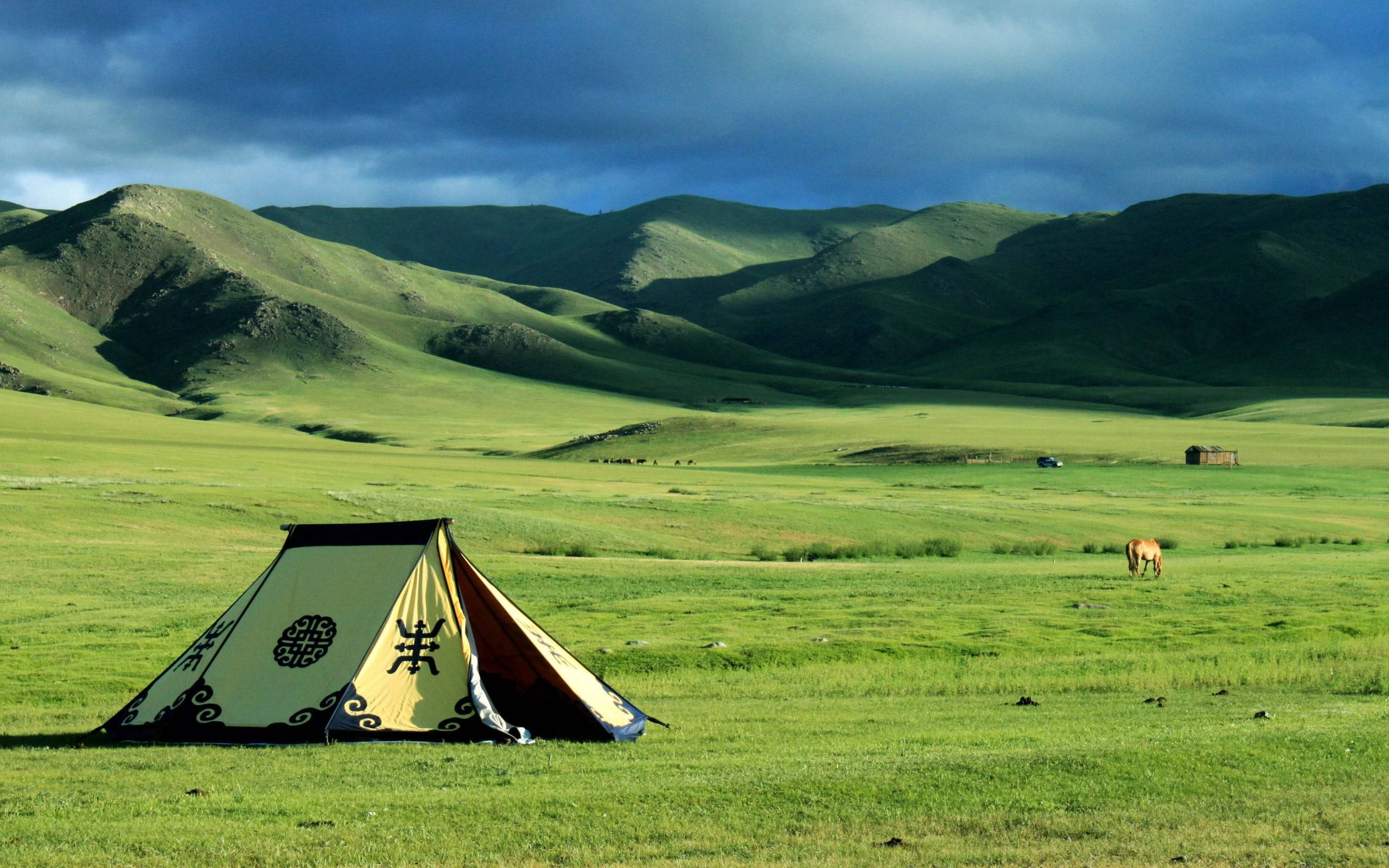 Mongolia 桌布 壁紙 — 一個色彩繽紛的傳統帳篷矗立在廣闊的綠色草地上，上方是戲劇性的暴風雲，遠處有連綿起伏的山丘和一匹正在吃草的馬。