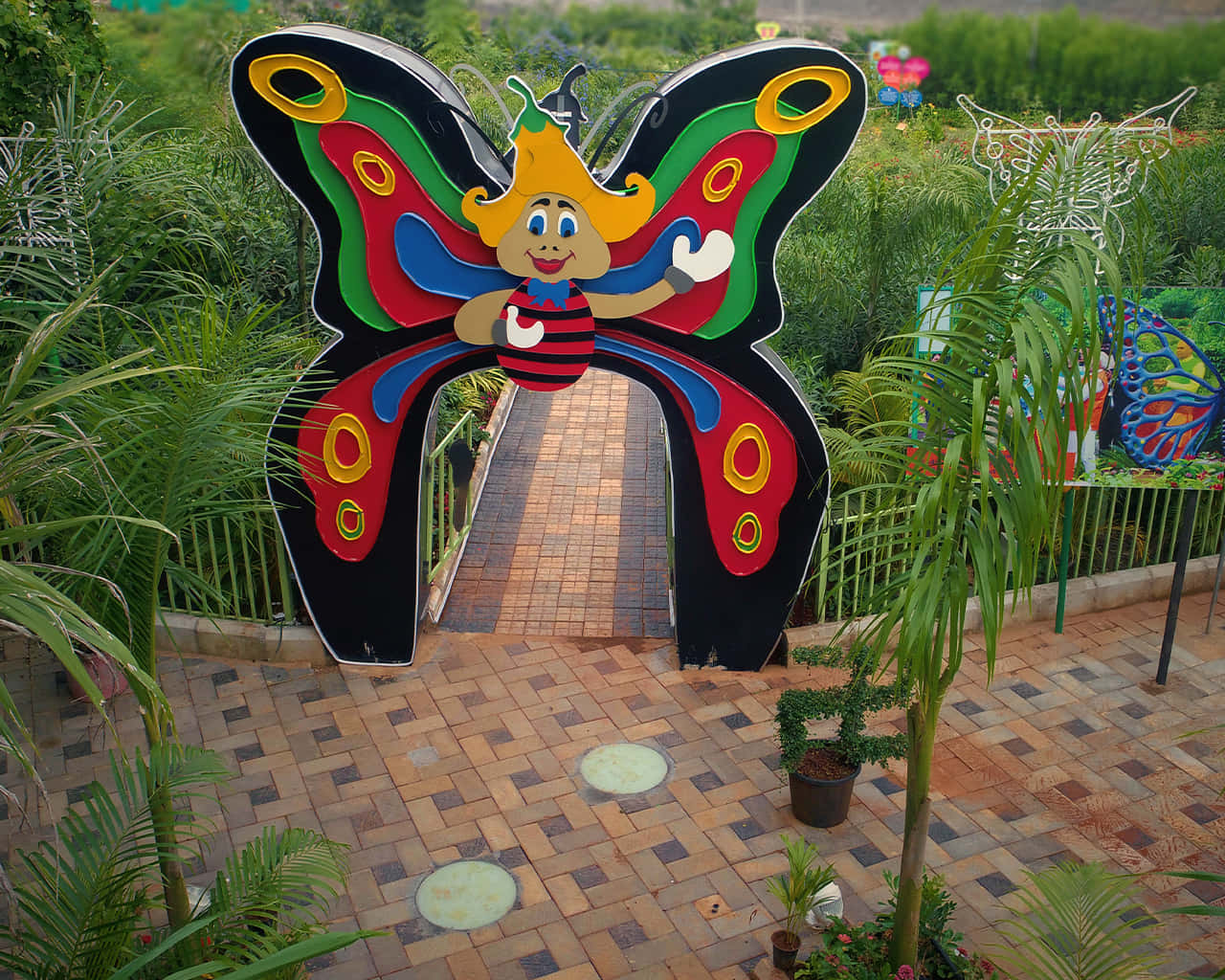 Butterfly Park 桌布 壁紙 — 一個色彩繽紛的卡通蝴蝶拱門，帶有微笑的臉，歡迎遊客穿過茂密的綠色花園小徑。