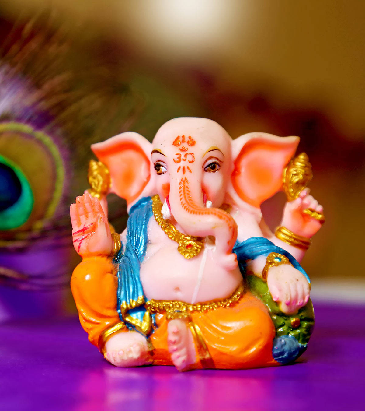 cute ganesha 桌布 壁紙 — 一個色彩繽紛的塑膠象神像盤腿坐在紫色表面上，背景有一根模糊的孔雀羽毛。