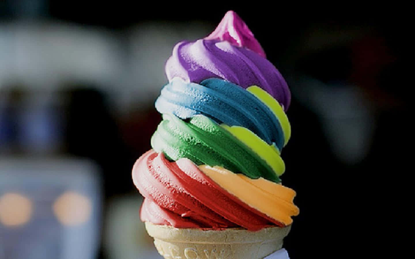 Ice Cream 桌布 壁紙 — 一個色彩繽紛的彩虹漩渦軟冰淇淋甜筒，鮮豔的顏色螺旋堆疊在深色模糊的背景上。