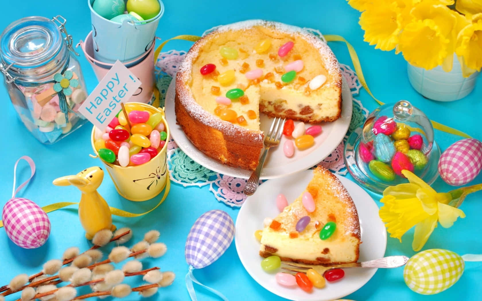 Spring Desserts 桌布 壁紙 — 一個色彩繽紛的復活節蛋糕，上面鋪滿了彩色糖果，放在明亮的藍色表面上，周圍環繞著水仙花、圖案蛋和裝滿糖果的罐子。