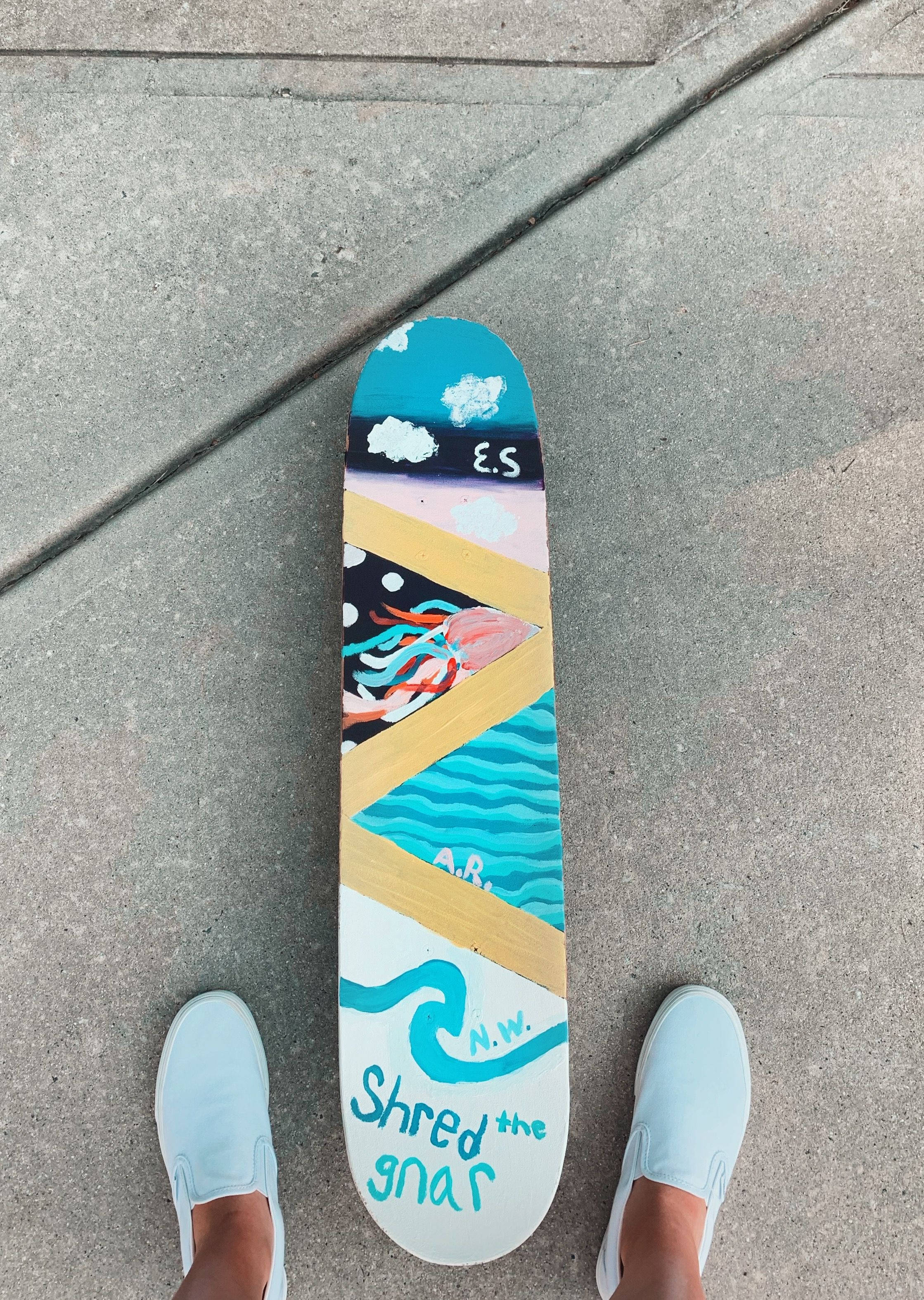 Skater Boy Aesthetic 桌布 壁紙 — 一個色彩繽紛的手繪滑板，上面有海浪和水母藝術，放在混凝土上，旁邊是淺藍色懶人鞋。