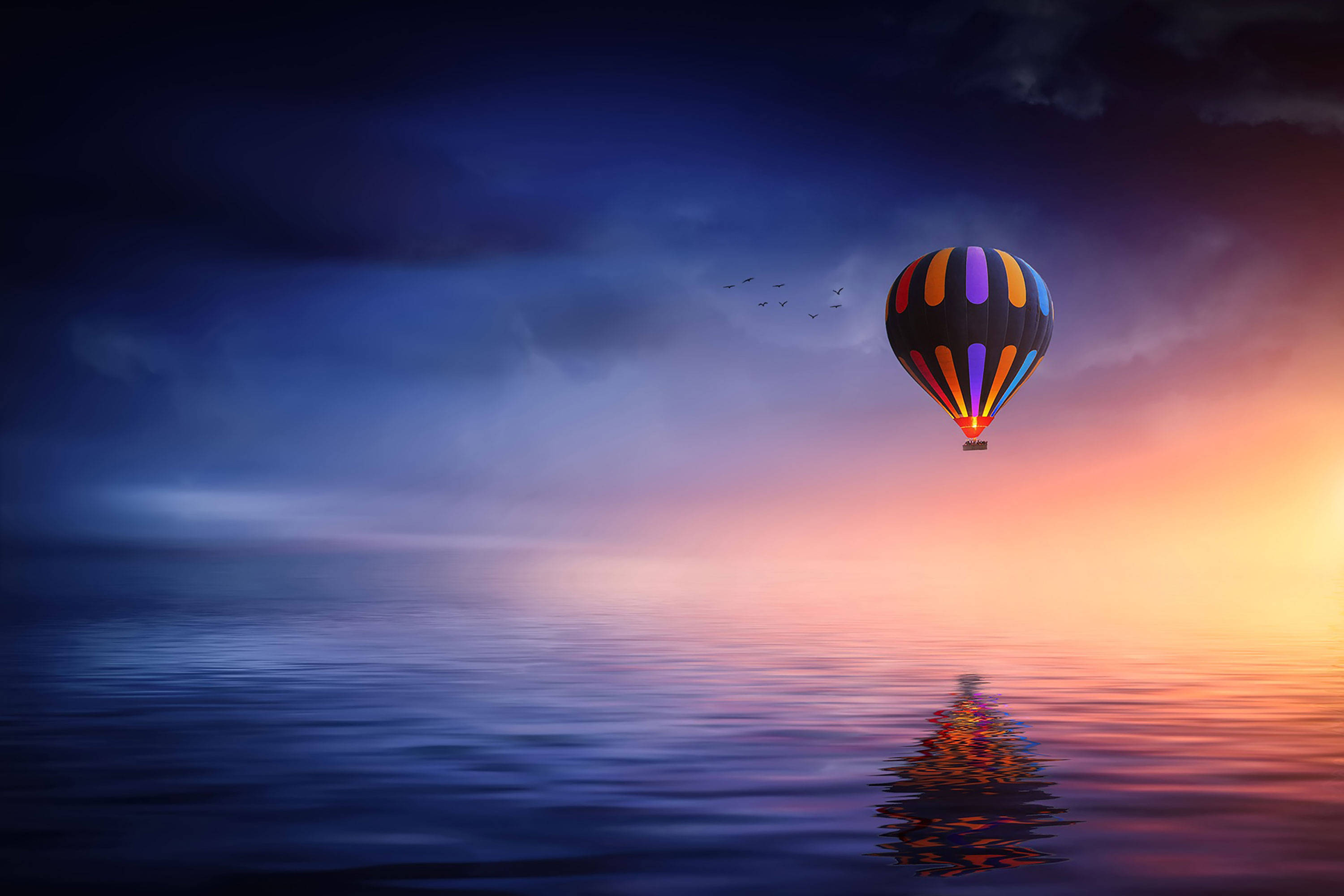 Hot Air Balloon 桌布 壁紙 — 一個色彩繽紛的熱氣球在日落時分漂浮在平靜的水面上，映出多彩的倒影，頭頂有鳥兒飛過。