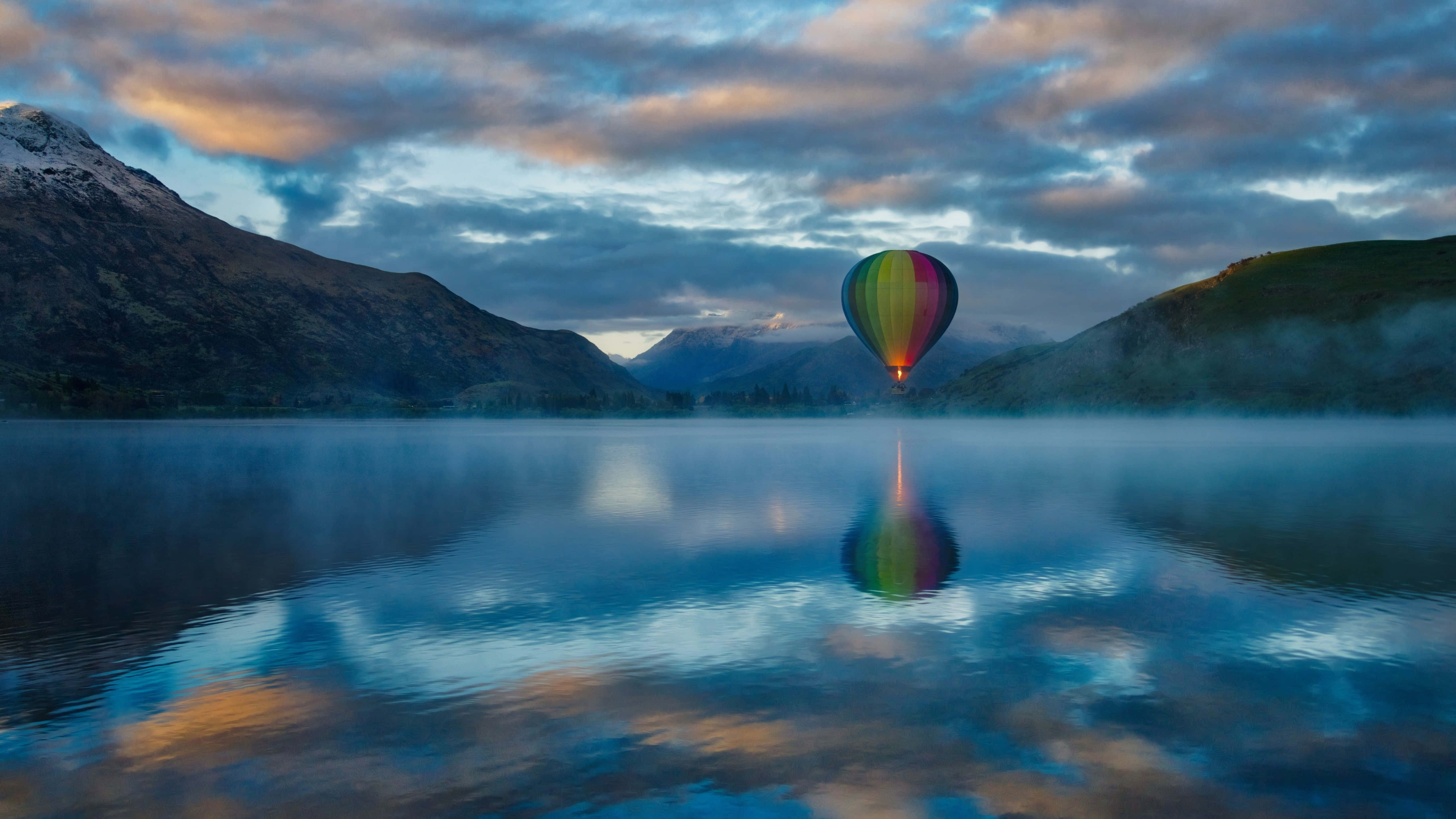 Hot Air Balloon 桌布 壁紙 — 一個色彩繽紛的熱氣球在黎明時分飄浮在霧氣瀰漫的湖面上，積雪的山峰倒映在平靜的水中。