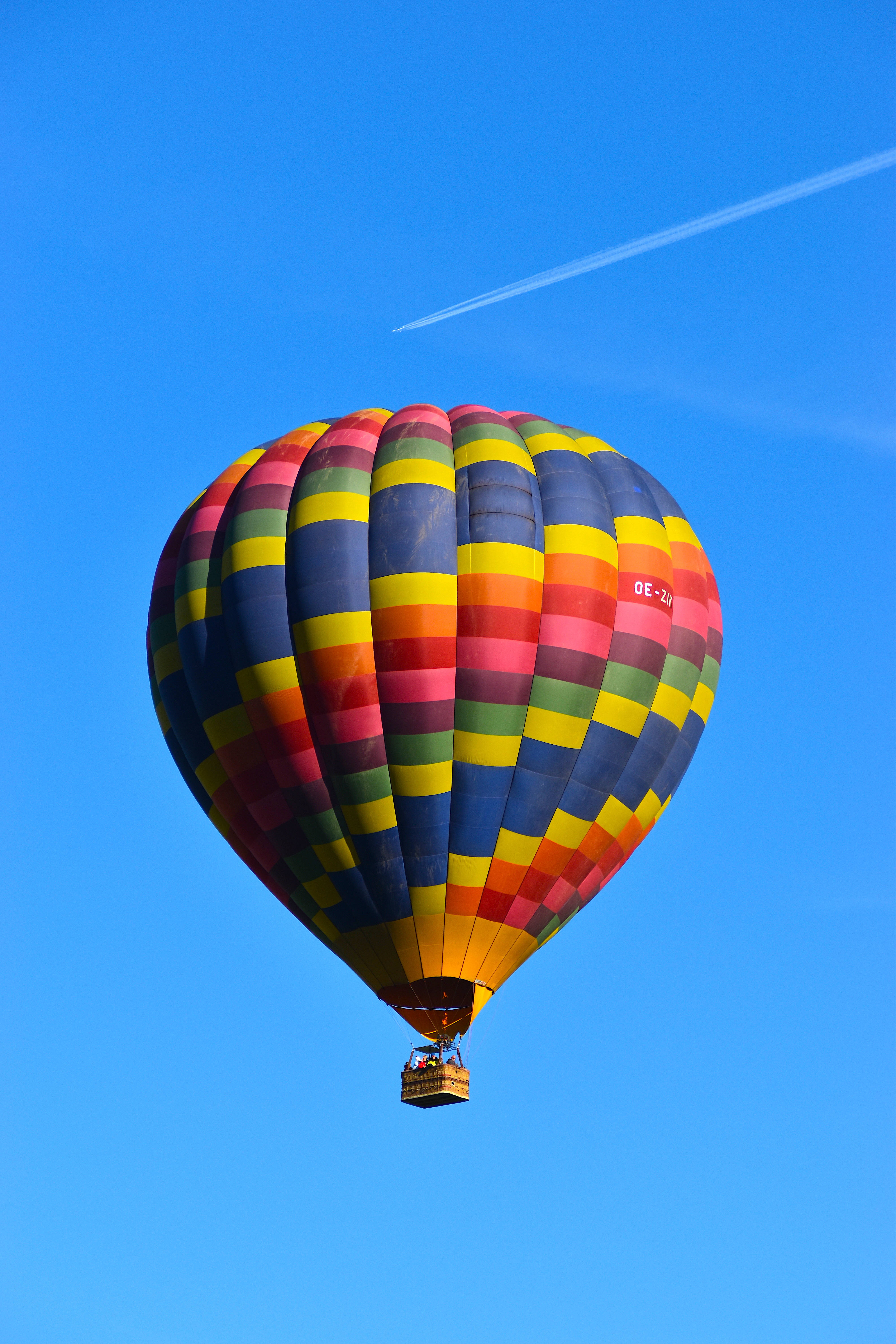 Balloon 桌布 壁紙 — 一個色彩繽紛的熱氣球帶有彩虹條紋，在晴朗的藍天中寧靜地漂浮，遠處有飛機的凝結尾跡。