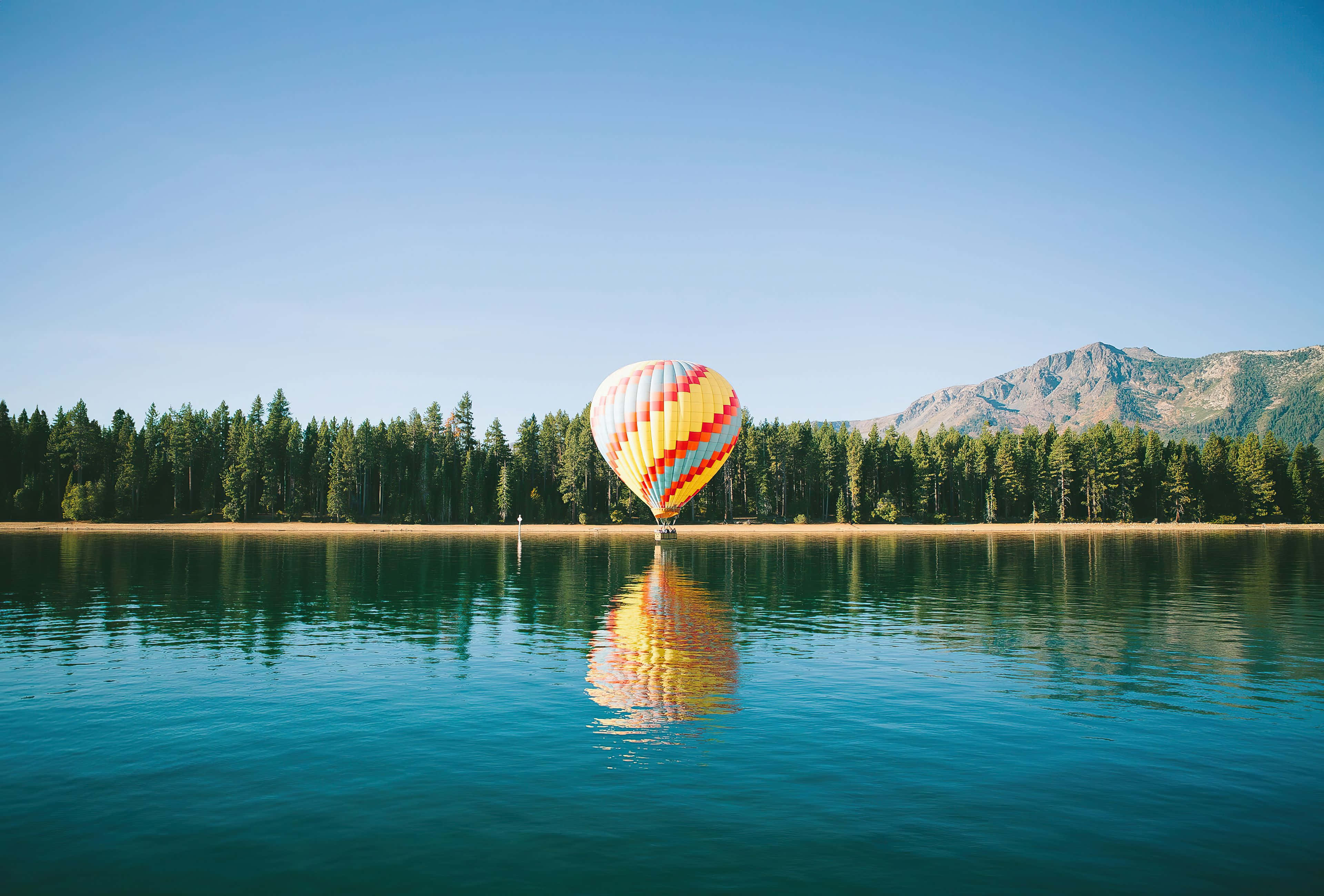 Hot Air Balloon 桌布 壁紙 — 一個色彩繽紛的熱氣球漂浮在平靜的藍綠色湖面上，湖岸樹木繁茂，遠處群山連綿，天空湛藍清澈。