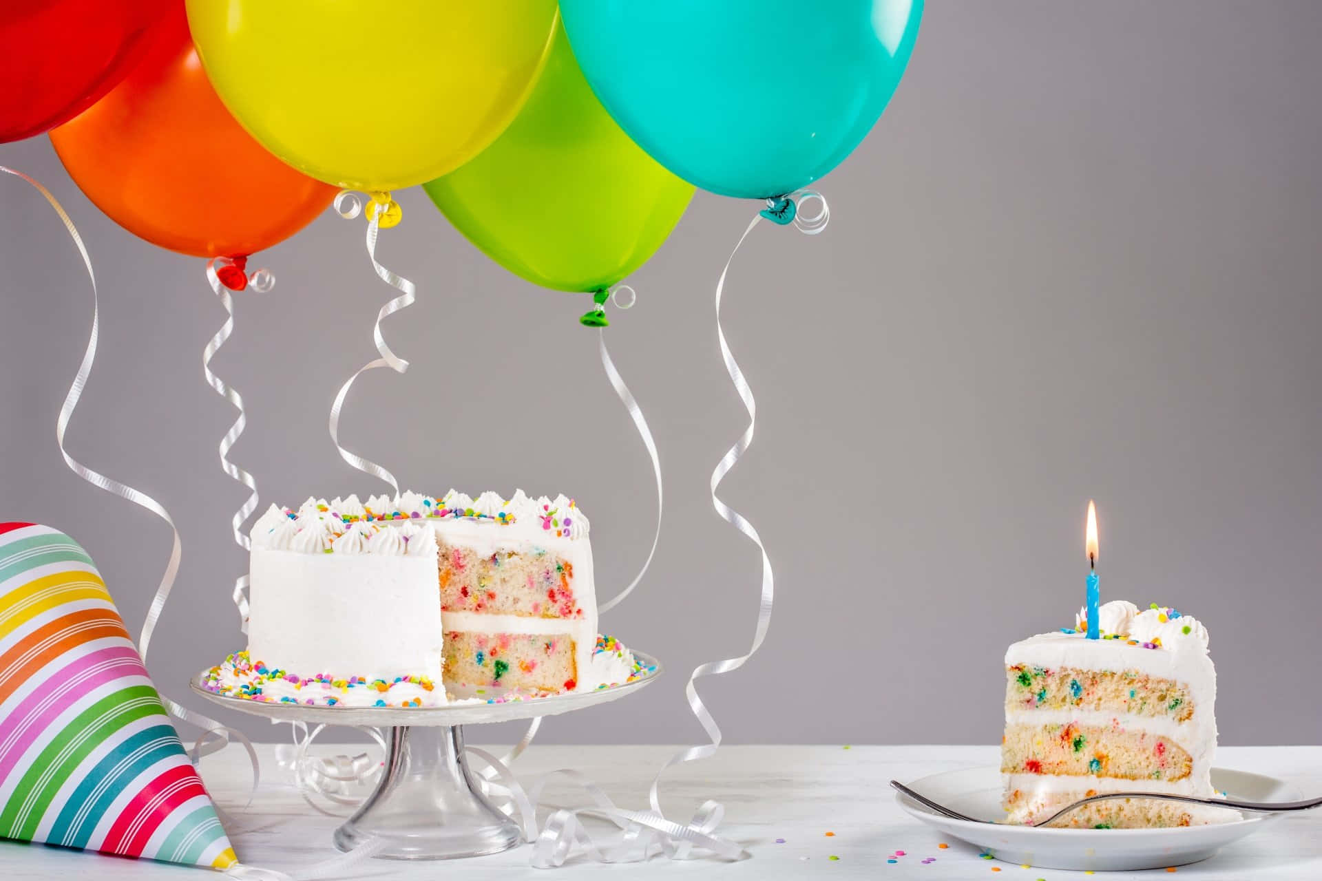 Birthday Cake 桌布 壁紙 — 一個色彩繽紛的生日蛋糕，上面有彩虹糖針，旁邊放著一塊點燃蠟燭的蛋糕，下方有明亮的氣球和彩帶。