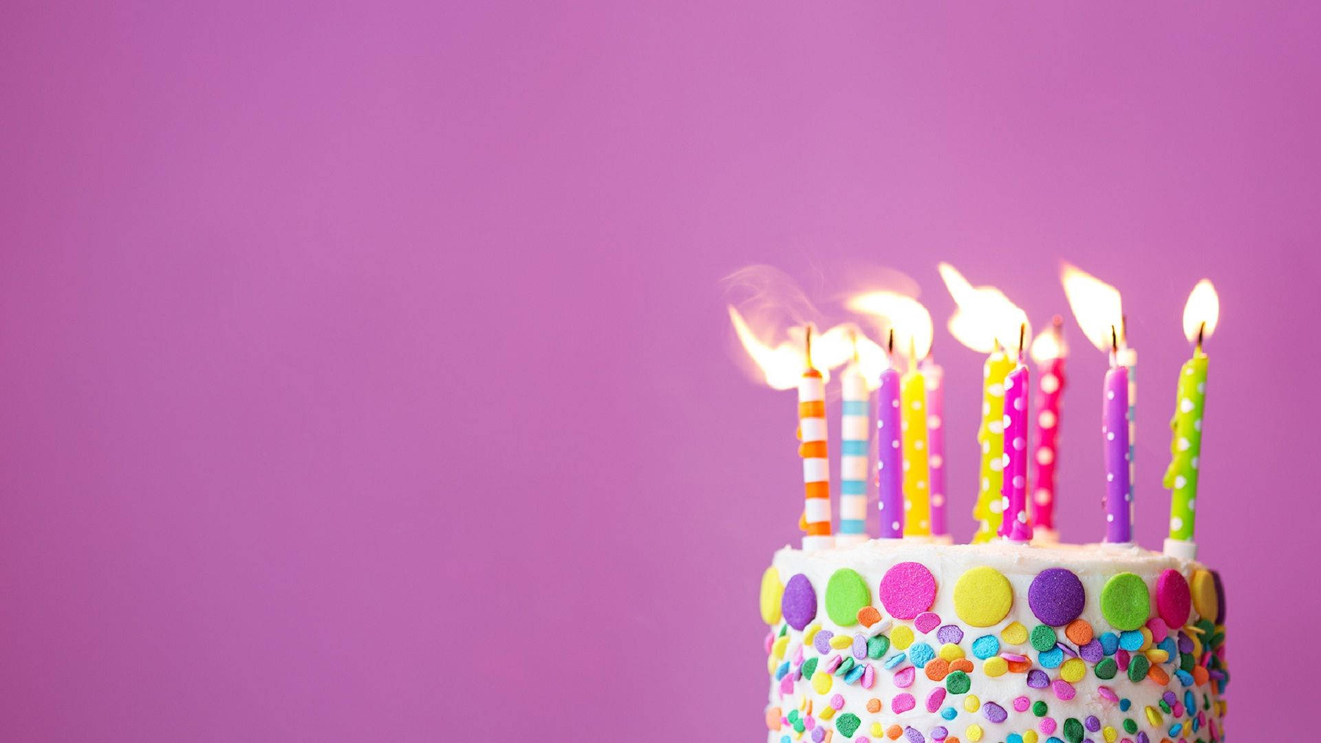 Birthday Cake 桌布 壁紙 — 一個色彩繽紛的生日蛋糕，上面有燃燒的蠟燭和彩虹糖針，背景為純紫色。