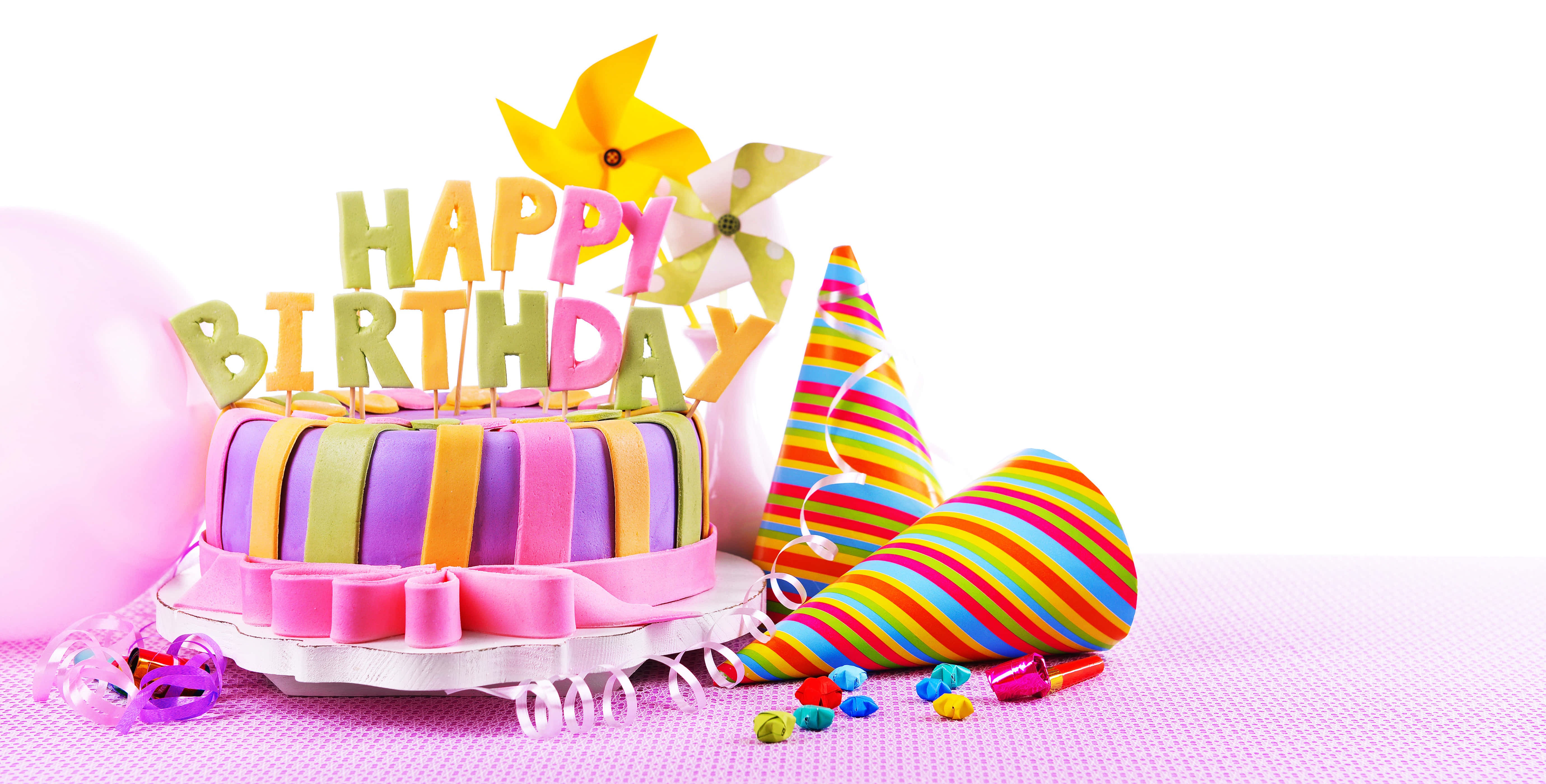 生日快樂 桌布 壁紙 — 一個色彩繽紛的生日蛋糕，帶有彩虹條紋和「HAPPY BIRTHDAY」裝飾，周圍環繞著派對帽和氣球，放在粉紅色表面上。