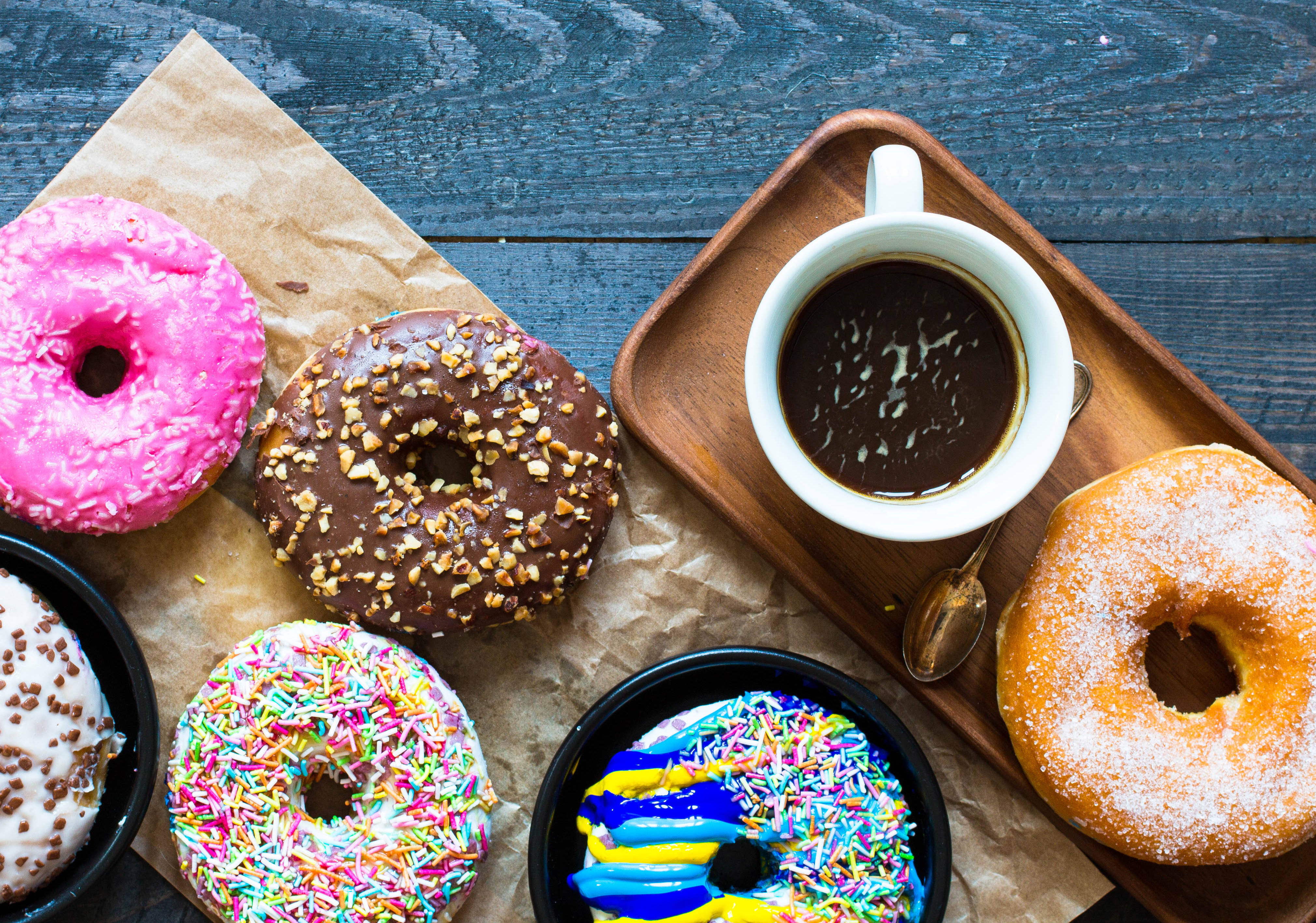 Donut 桌布 壁紙 — 一個色彩繽紛的組合，包括釉面和裝飾的甜甜圈，排列在烘焙紙上，旁邊有一杯咖啡在木托盤上。