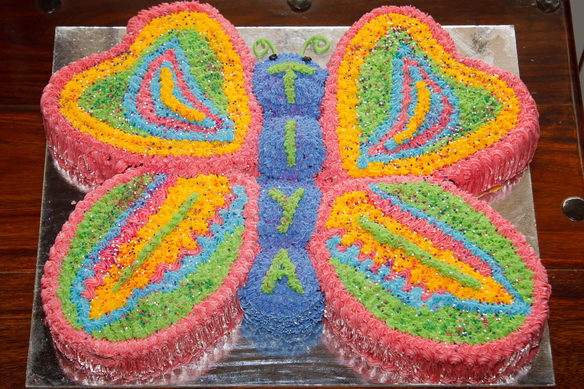 Butterfly Cake 桌布 壁紙 — 一個色彩繽紛的蝴蝶形蛋糕，帶有彩虹色糖霜和彩色糖粒，放在木桌上的銀托盤上。