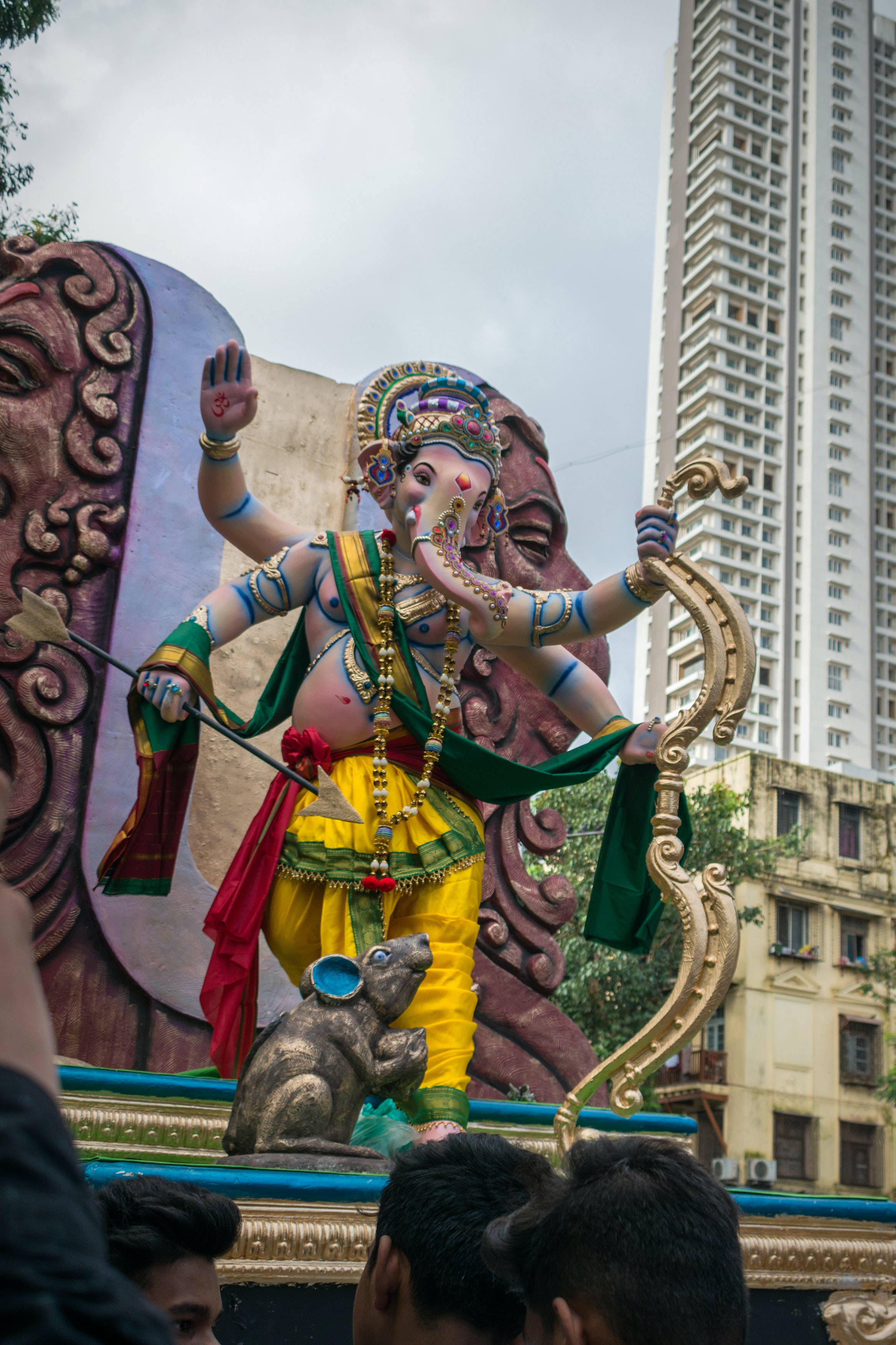 Ganesh 桌布 壁紙 — 一個色彩繽紛的象頭神像，擁有象頭和人身，站在節日裝飾的花車上，背景是現代城市的高樓大廈。