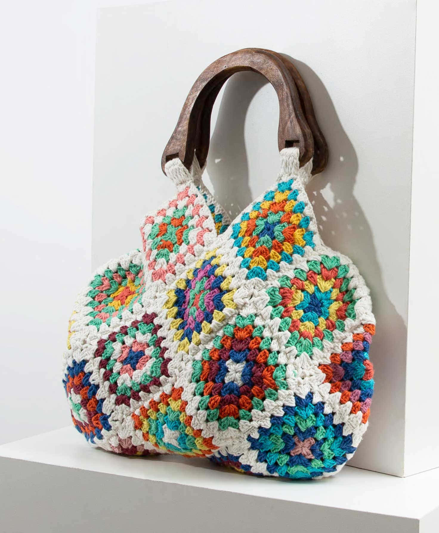 Crocheting 桌布 壁紙 — 一個色彩繽紛的鉤針編織祖母方格手提袋，配有木製手柄，放在白色架子上，背景是純白牆壁。