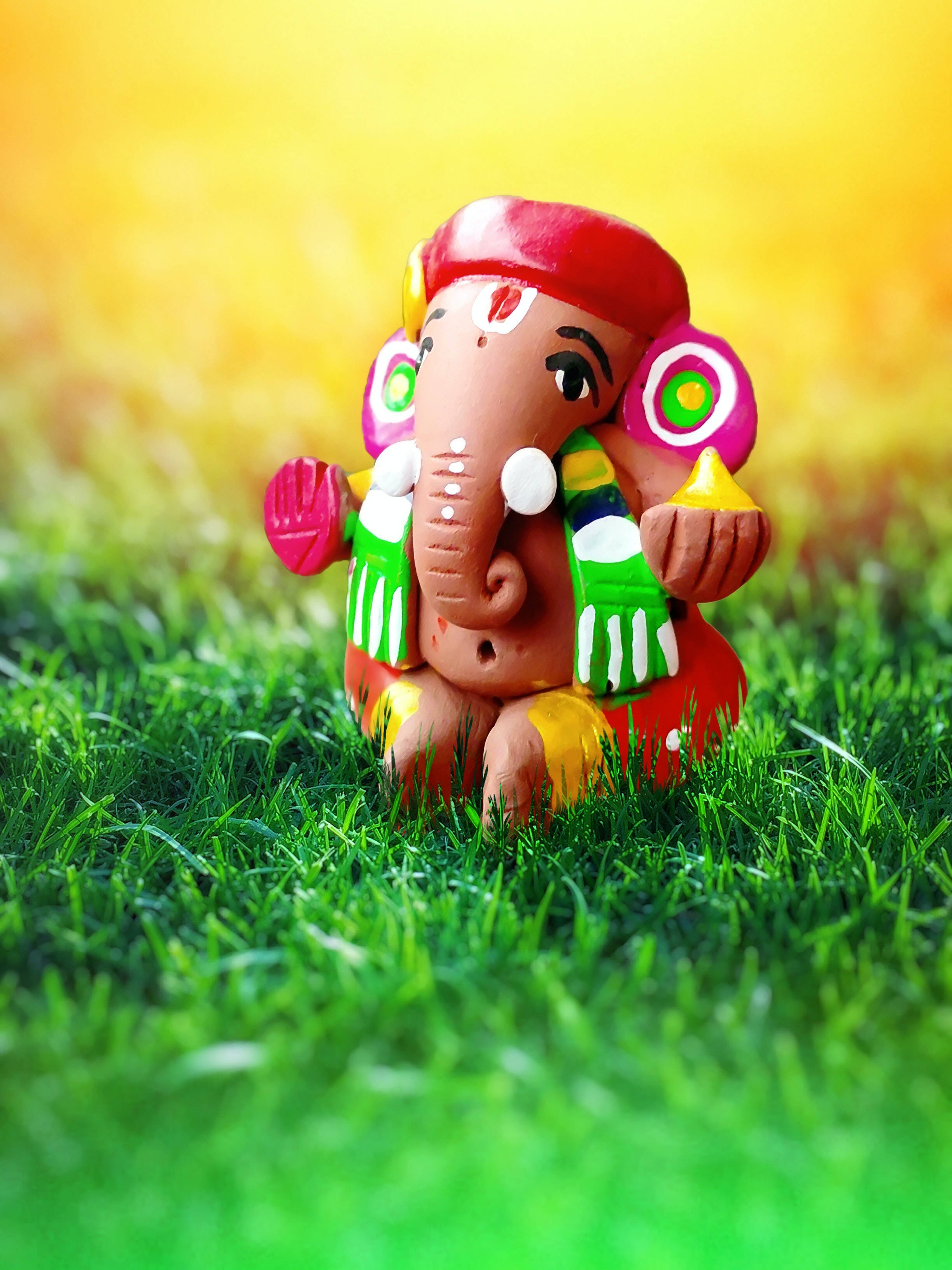 ganesh iPhone 桌布 壁紙 — 一個色彩鮮豔的彩繪黏土象頭神雕像，在溫暖的金色天空下，寧靜地坐在茂密的綠草地上。